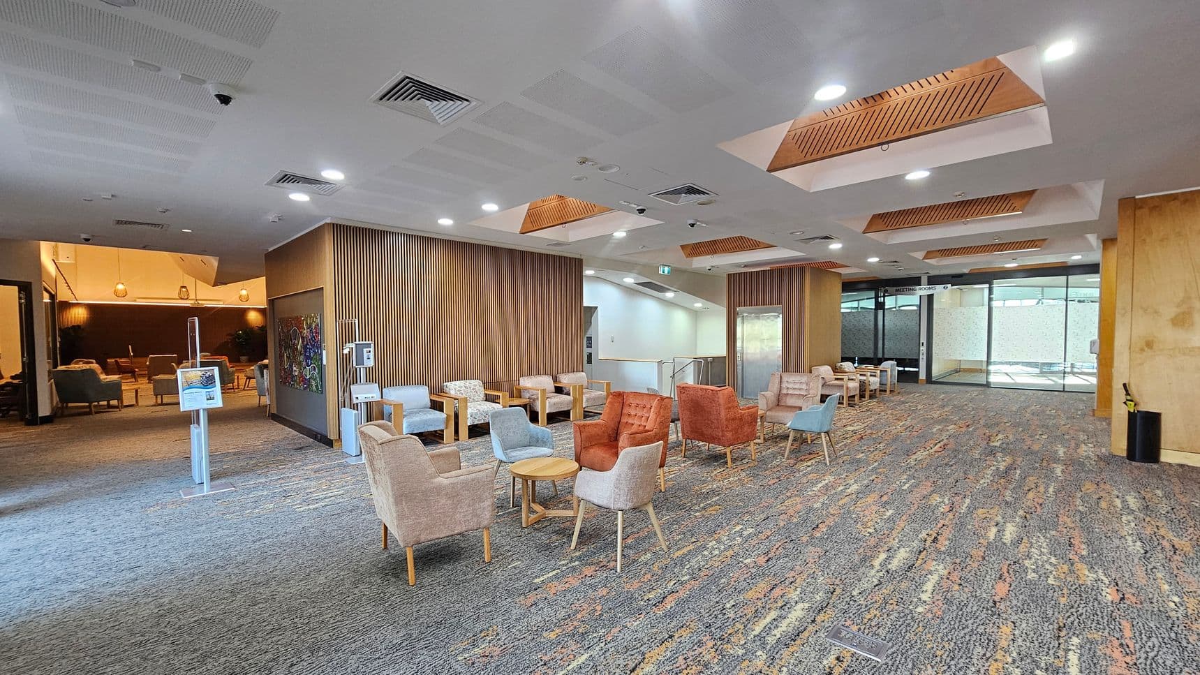 A spacious lobby area with a patterned carpet floor. On the left, there is a seating area with various chairs and small tables. The middle features a wooden panelled wall with a colourful artwork. On the right, more seating is arranged, and a glass door labelled "Meeting Rooms" is visible. The ceiling has recessed lighting and wooden accents. The background shows another seating area with warm lighting and pendant lamps.