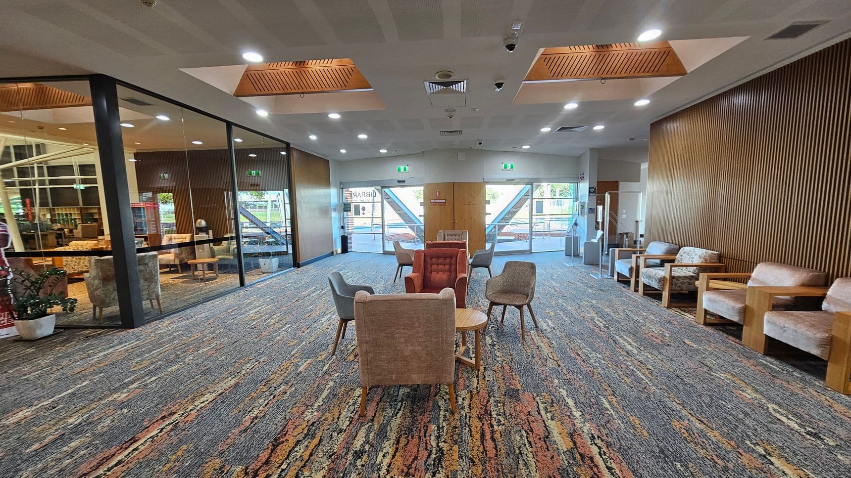 A spacious room with a patterned carpet in shades of grey, orange, and brown. In the centre, there are several chairs and small tables arranged in a casual seating area. The left side features a glass wall with a view into another room, while the right side has a wooden panelled wall with additional seating. The ceiling has recessed lighting and wooden accents. At the far end, there are large glass doors leading outside, flanked by emergency exit signs.