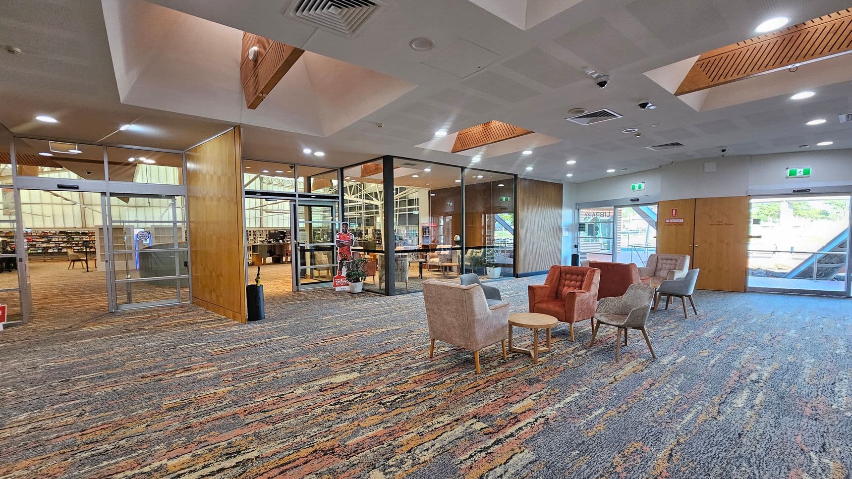 A spacious interior with a patterned carpet floor. On the right, there are several upholstered chairs and small round tables arranged in a seating area. The left side features glass doors leading to a library with visible bookshelves. The ceiling has recessed lighting and wooden accents. A cardboard cutout and potted plants are near the entrance. Large windows and doors provide natural light from outside.