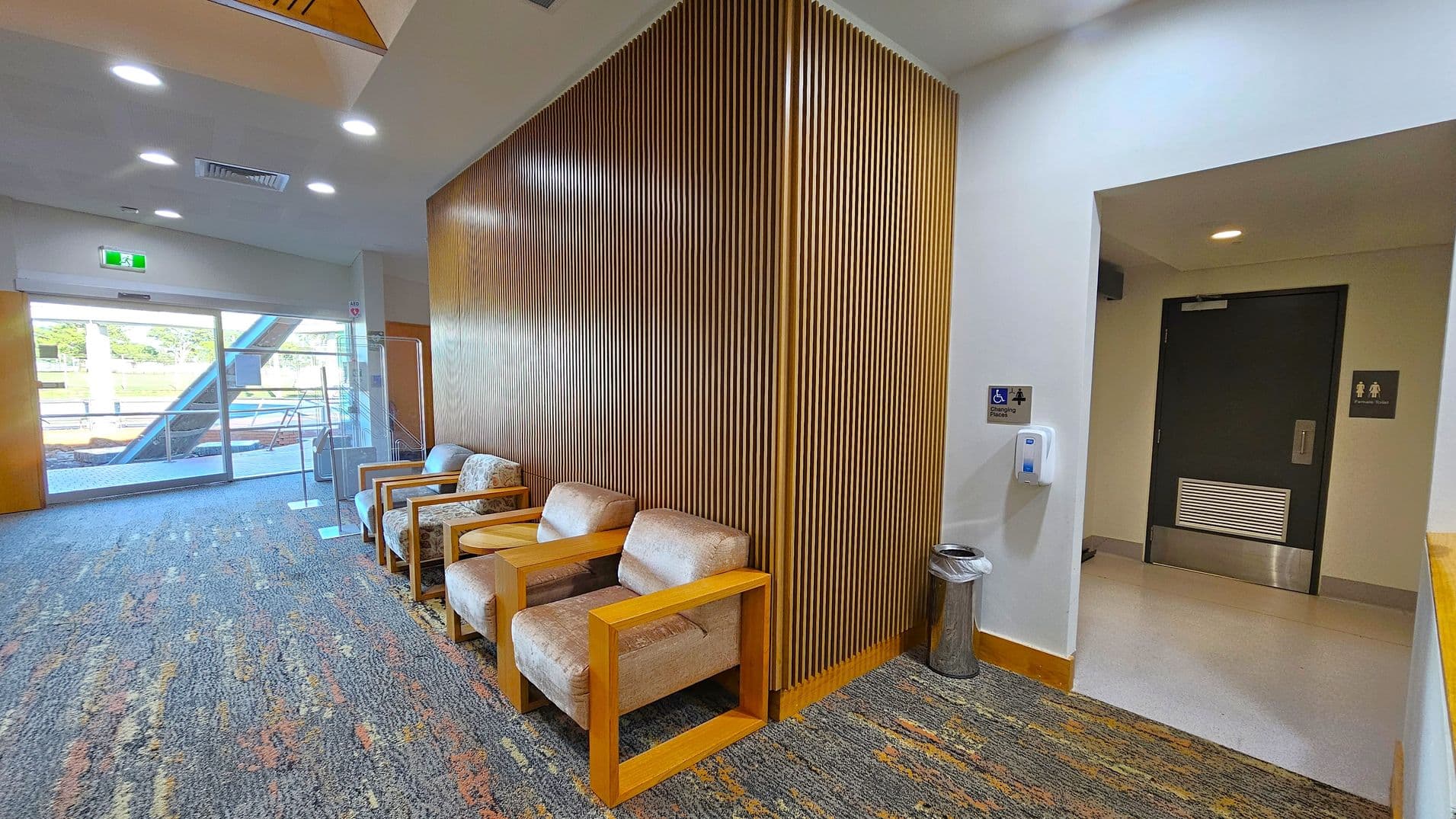 On the left, there is a glass door leading outside, with a green exit sign above. The floor is carpeted with a multicoloured pattern. In the middle, there are several cushioned chairs with wooden armrests against a wall with vertical wooden slats. On the right, a hallway leads to a dark door with restroom signs. A hand sanitiser dispenser and a small bin are mounted on the wall. The ceiling is white with recessed lighting.