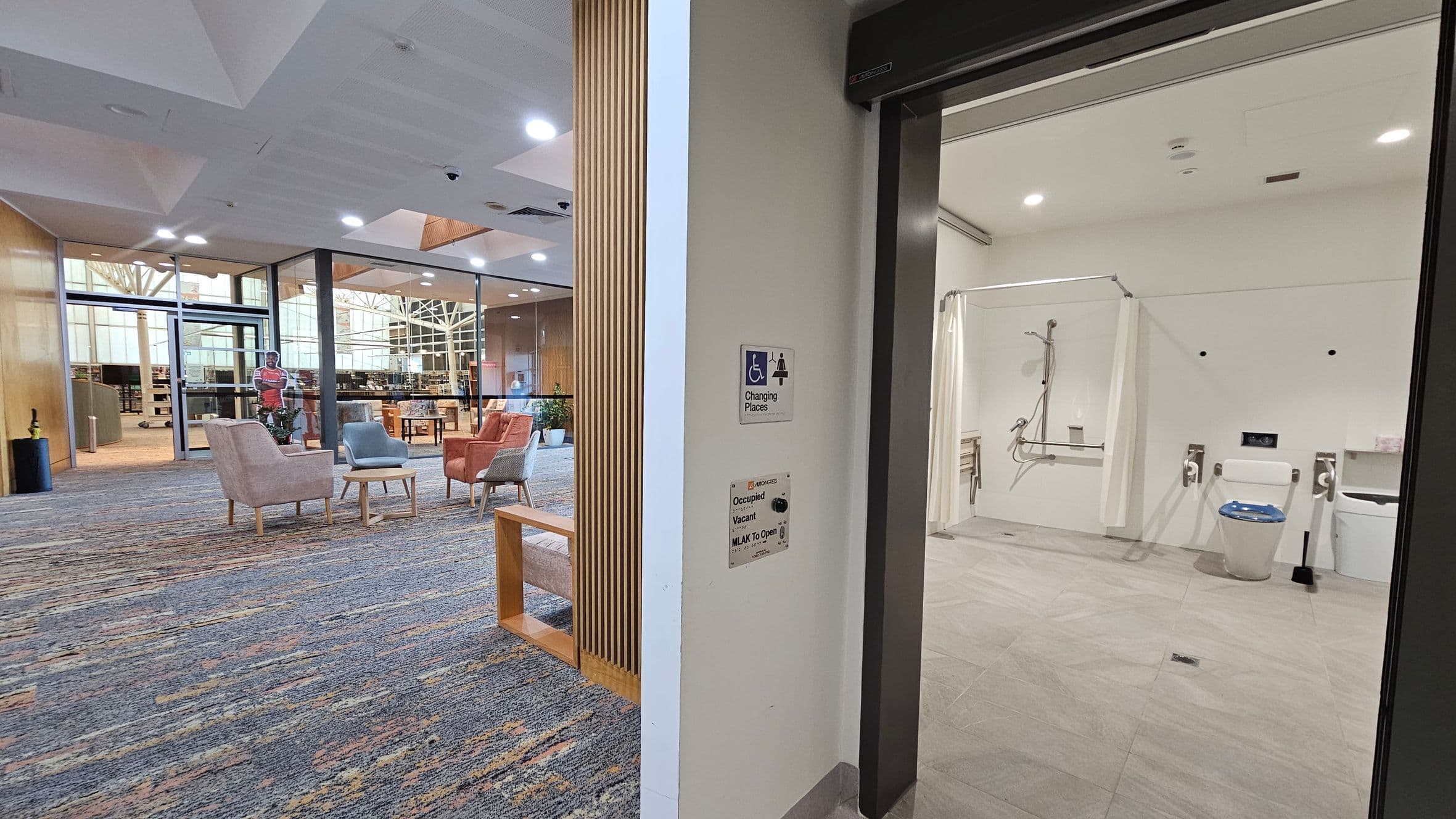 On the right, there's an accessible bathroom with a shower area, toilet, and support rails. The floor is tiled, and the ceiling has recessed lighting. On the left, a seating area with pastel-coloured chairs and small tables is visible, set on a patterned carpet. Beyond, glass walls reveal a library space with bookshelves and a cut-out figure. The ceiling is white with embedded lights, and a wooden partition partially separates the two areas.