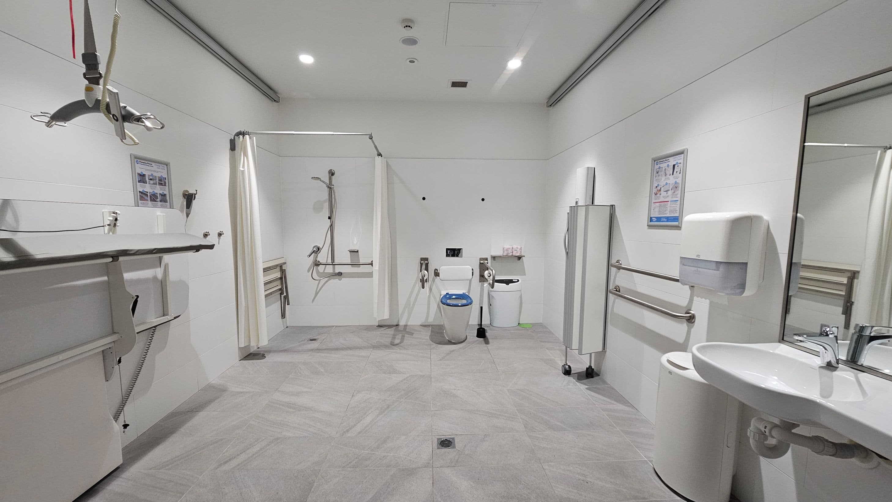 A spacious, accessible bathroom with grey tiled flooring and white walls. On the left, there is a height-adjustable changing table and a ceiling hoist. In the centre, a shower area with a curtain and a toilet equipped with support rails. On the right, a sink with a mirror above, a soap dispenser, and a waste bin. The ceiling is fitted with recessed lighting. Various informational posters are mounted on the walls.