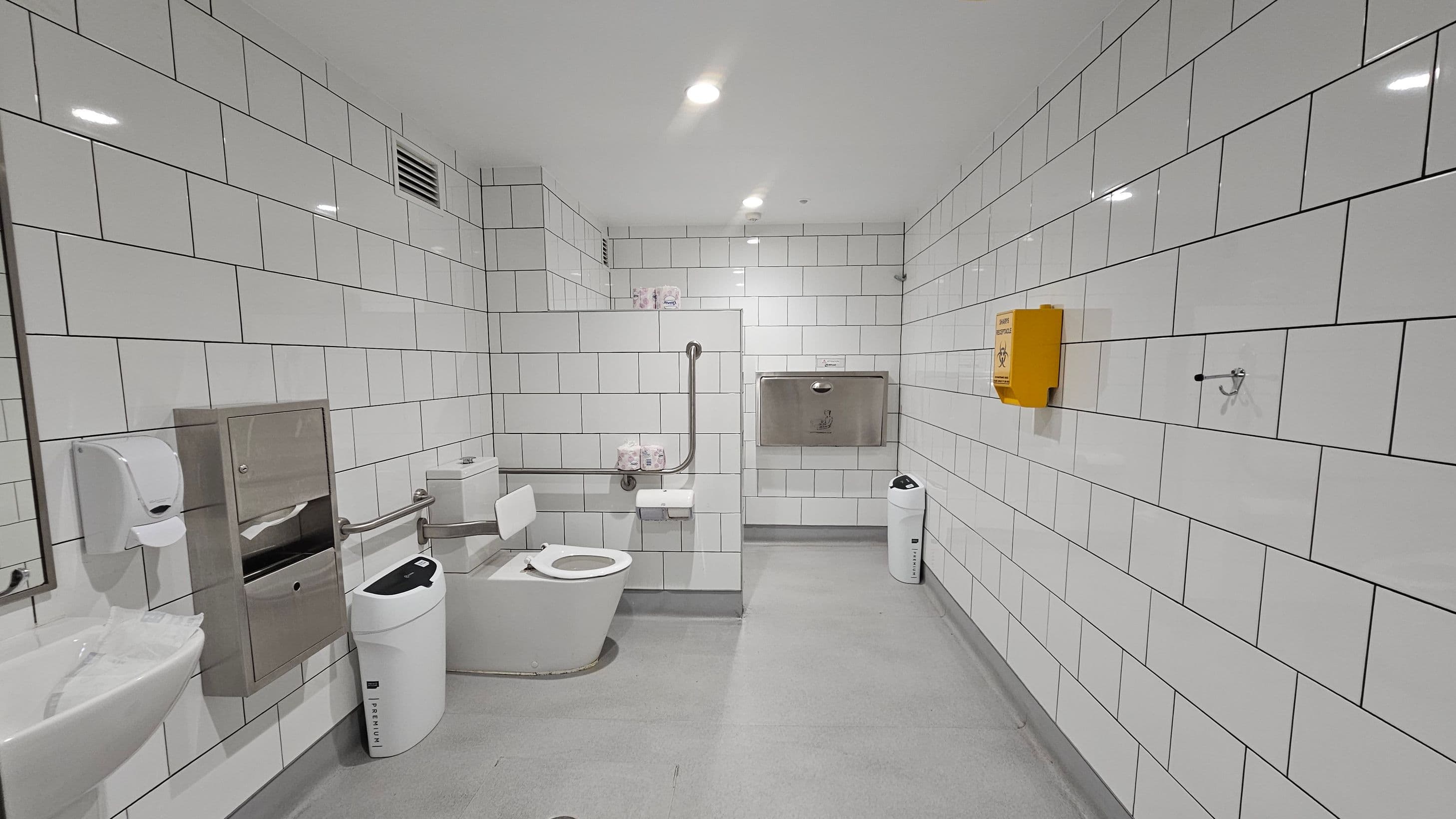 A clean, accessible bathroom with white tiled walls and a light grey floor. On the left, there's a sink with a soap dispenser and a paper towel holder. A toilet with support rails is positioned centrally, with toilet paper and a sanitary bin nearby. On the right, a yellow biohazard disposal box is mounted on the wall. The ceiling is white with recessed lighting, and a baby changing station is visible on the back wall.