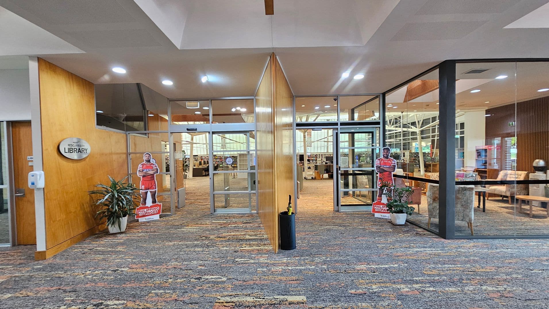 Entrance to a library with a glass door in the centre, flanked by wooden panels. On the left, a sign reads "Library" above a sanitiser dispenser and a potted plant. On the right, a similar potted plant and a cardboard cut-out of a person in a red outfit. Inside, bookshelves and seating areas are visible. The floor is carpeted with a multicoloured pattern, and the ceiling has recessed lighting.