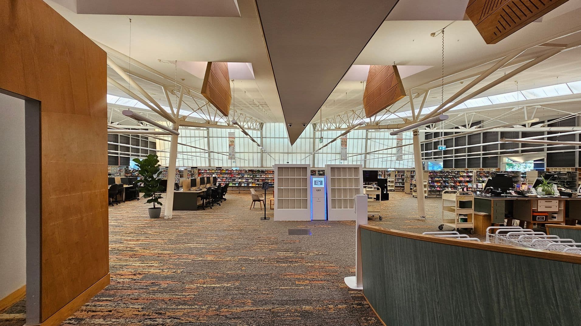 A spacious library interior with a high ceiling featuring exposed beams and large windows in the background. In the centre, there are two white shelving units with a digital kiosk between them. To the left, there are rows of computer stations and a potted plant. On the right, a curved desk with baskets and office supplies. The floor is covered with a patterned carpet in earthy tones. The ceiling has suspended wooden panels and modern lighting fixtures.