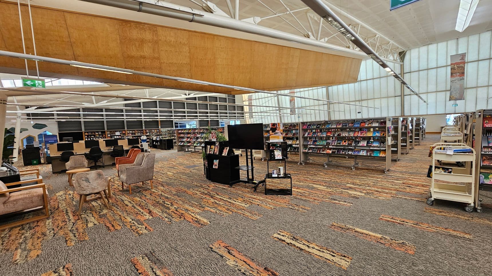 A spacious library interior with a patterned carpet floor. On the left, there are several comfortable chairs and computer stations with black chairs. In the centre, a black stand holds a large screen and some books. To the right, rows of bookshelves are filled with books, and there are book carts nearby. The ceiling is high with exposed beams and a large wooden panel. The walls are made of translucent panels, allowing natural light to filter through.