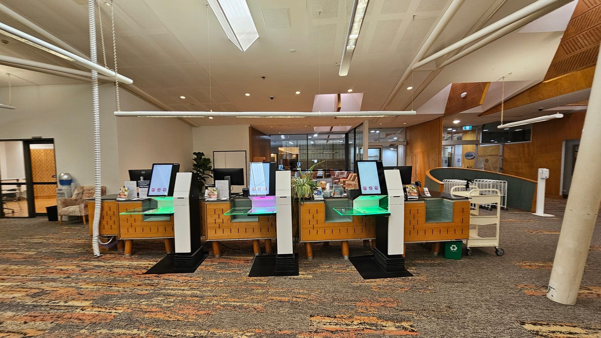 A library interior with a row of self-service kiosks in the centre, each with a screen and a glowing base. Behind them is a wooden counter with various items and a plant. To the left, there's a seating area with a water cooler and chairs. On the right, a book cart and a recycling bin are visible. The floor is carpeted with a multicoloured pattern, and the ceiling features white panels and lights. The background includes glass doors and wooden walls.