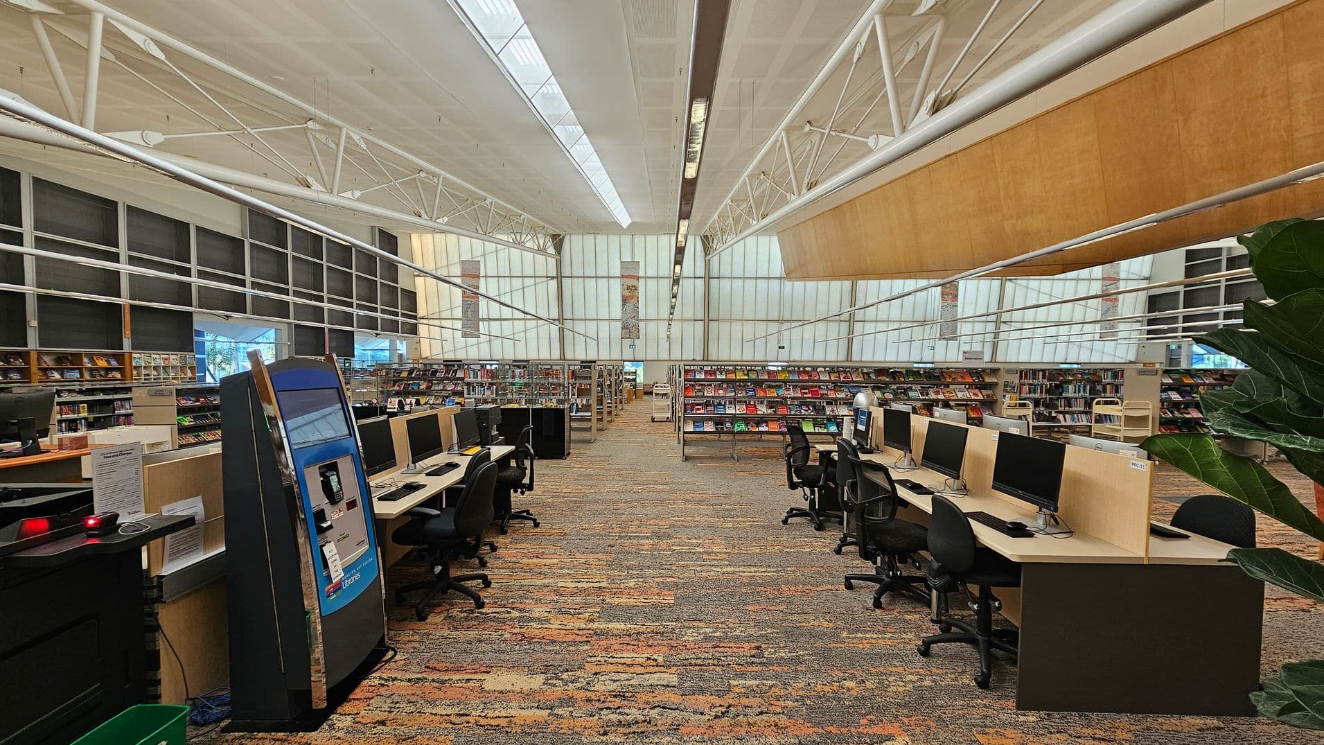 A spacious library interior with a high ceiling featuring exposed white beams. In the foreground, rows of computer stations with black chairs are on both sides. The floor is covered with a patterned carpet in earthy tones. In the background, bookshelves filled with colourful books line the walls. Large windows on the left allow natural light to illuminate the space. A large plant is partially visible on the right.