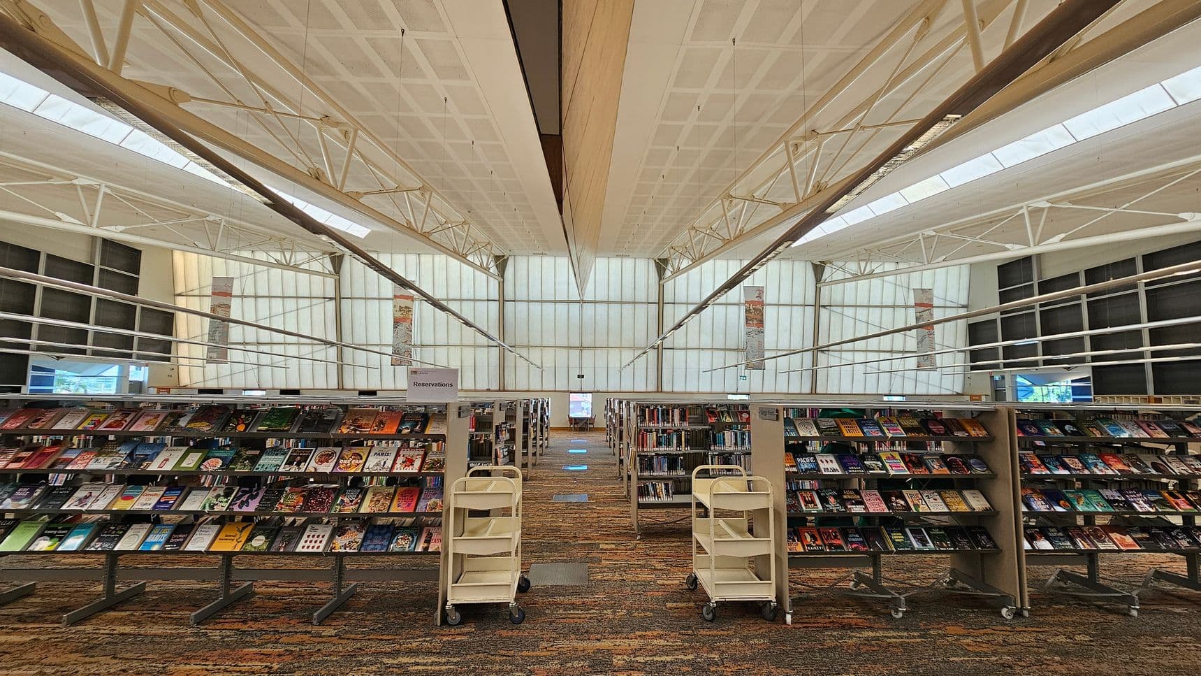 Bookshelves filled with various books are in the foreground, with two white book carts positioned in front. The background features large, translucent windows allowing natural light to illuminate the space. The ceiling is high with visible metal beams and a grid pattern. The floor is carpeted with a multicoloured pattern. The overall layout is spacious and organised, with a clear path between the shelves.
