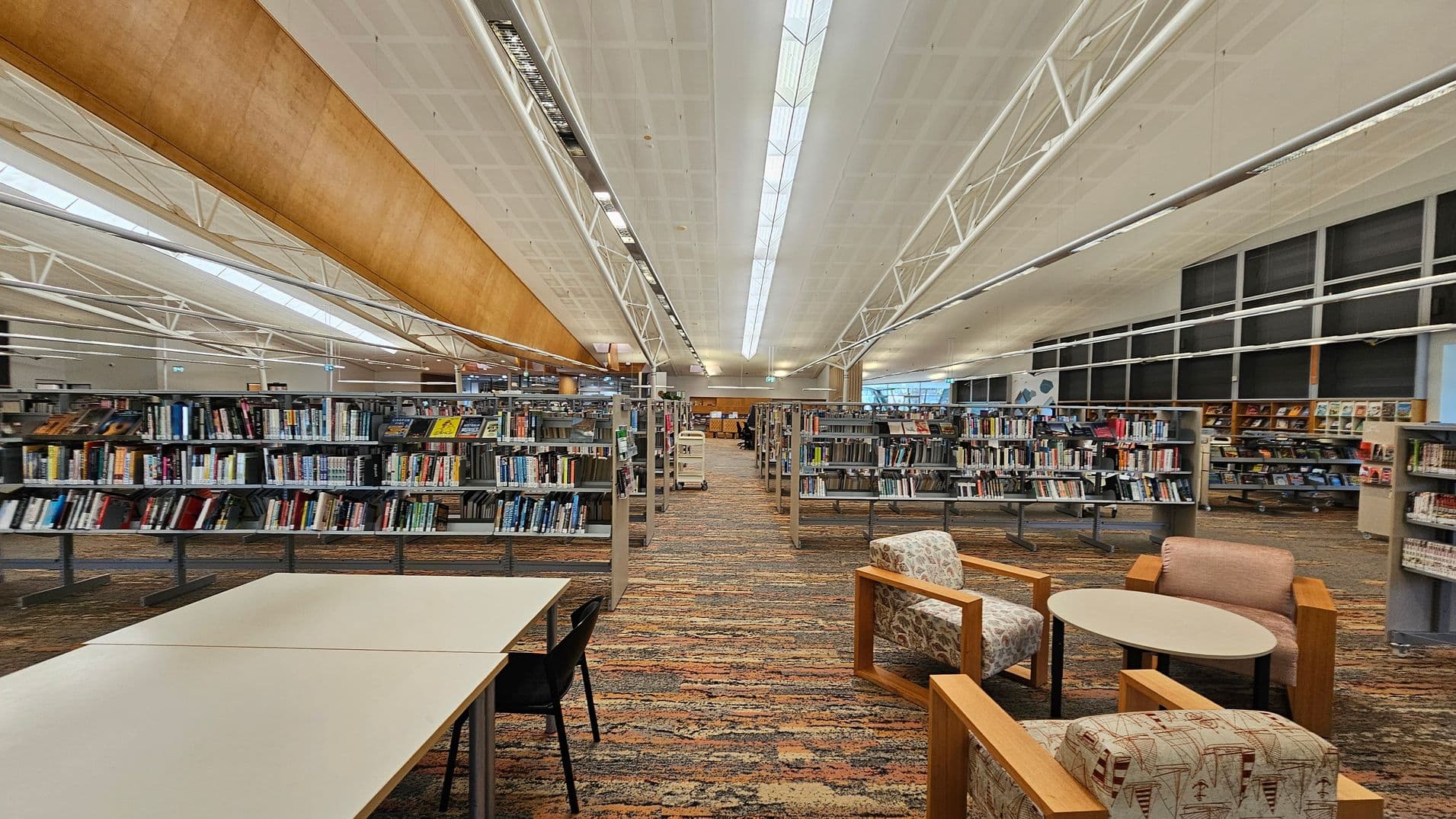 A spacious library interior with rows of bookshelves filled with books in the centre. On the left, there are white tables with black chairs. On the right, there are cushioned chairs with patterned upholstery and a small round table. The floor is covered with a multicoloured carpet. The ceiling is high with white panels and long fluorescent lights. Large windows are visible in the background, allowing natural light to enter.
