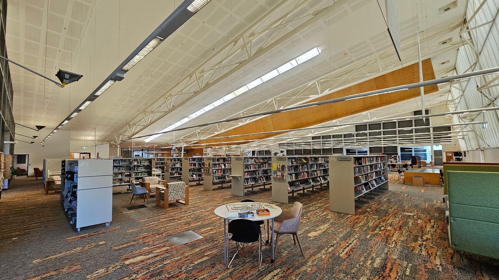 A spacious library interior with a high, white ceiling featuring exposed beams and skylights. In the foreground, a round table with chairs is placed on a multicoloured carpet. Bookshelves filled with books are arranged in rows across the room. On the left, there are more bookshelves and a few armchairs. The right side has additional seating and a green partition. The background shows large windows and more tables and chairs. The lighting is bright and evenly distributed.