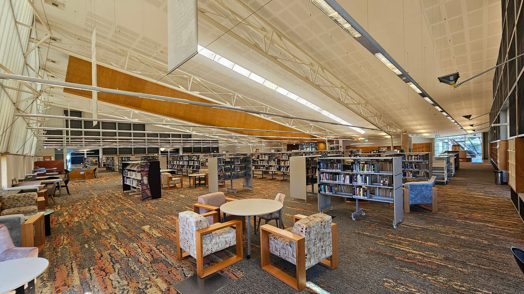 A spacious library interior with a high, white ceiling featuring exposed beams and skylights. In the foreground, there are round tables surrounded by patterned armchairs. The floor is covered with a multicoloured carpet. Bookshelves filled with books are arranged in rows across the middle and background. On the left, there are additional seating areas with tables and chairs. The right side has more bookshelves and a large window allowing natural light to enter.