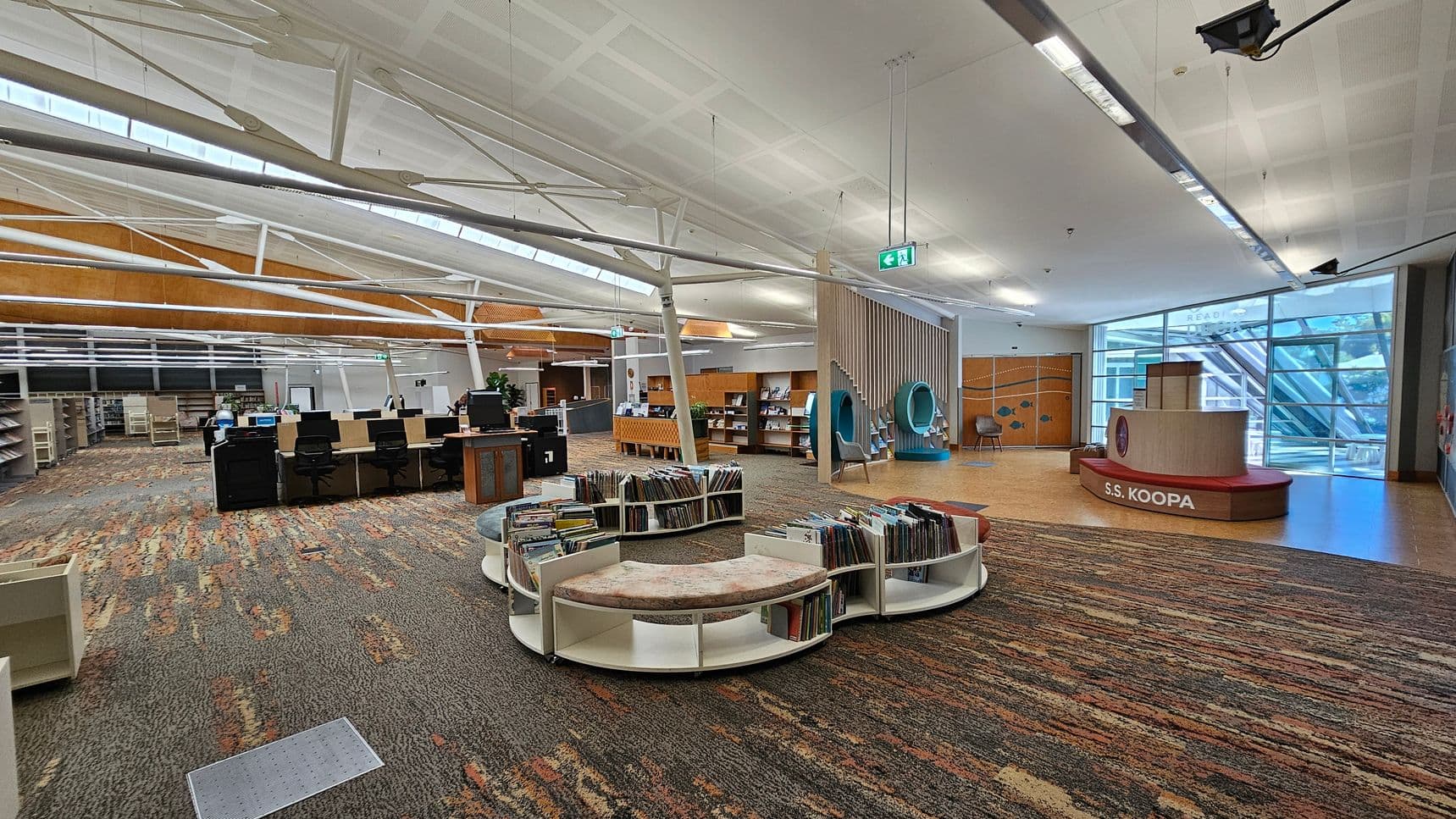 A spacious library interior with a colourful, patterned carpet. On the left, there are rows of bookshelves and a line of computer desks with chairs. In the centre, circular bookshelves with cushioned seating display books. On the right, a play area features a structure labelled "S.S. KOOPA" and two circular reading nooks. The ceiling is high with exposed beams, and large windows in the background allow natural light to enter.