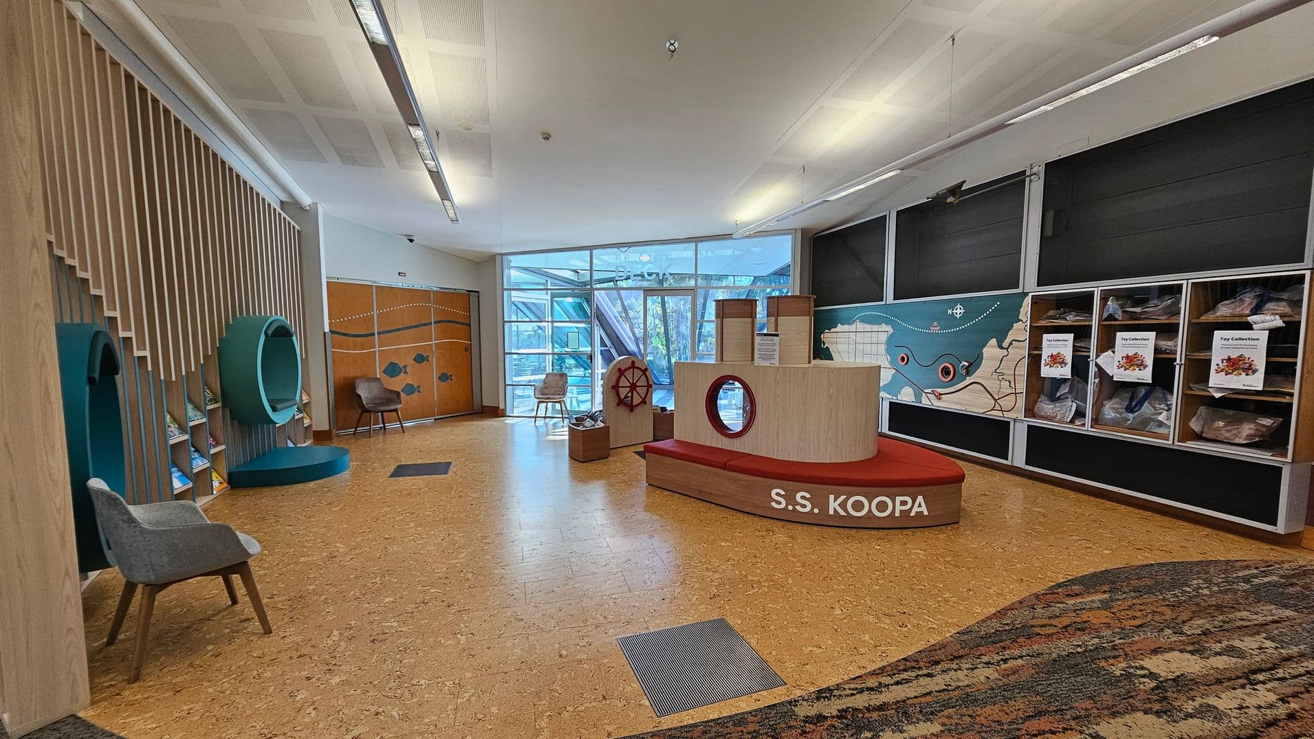 A spacious room with a cork floor and a white ceiling. On the left, there are wooden slats with circular green structures and a grey chair. In the centre, a ship-themed play area labelled "S.S. KOOPA" with a red bench and a ship's wheel. The right side features a wall with a map and display cases labelled "Toy Collection." The background has large windows allowing natural light, and a wooden door with fish designs.