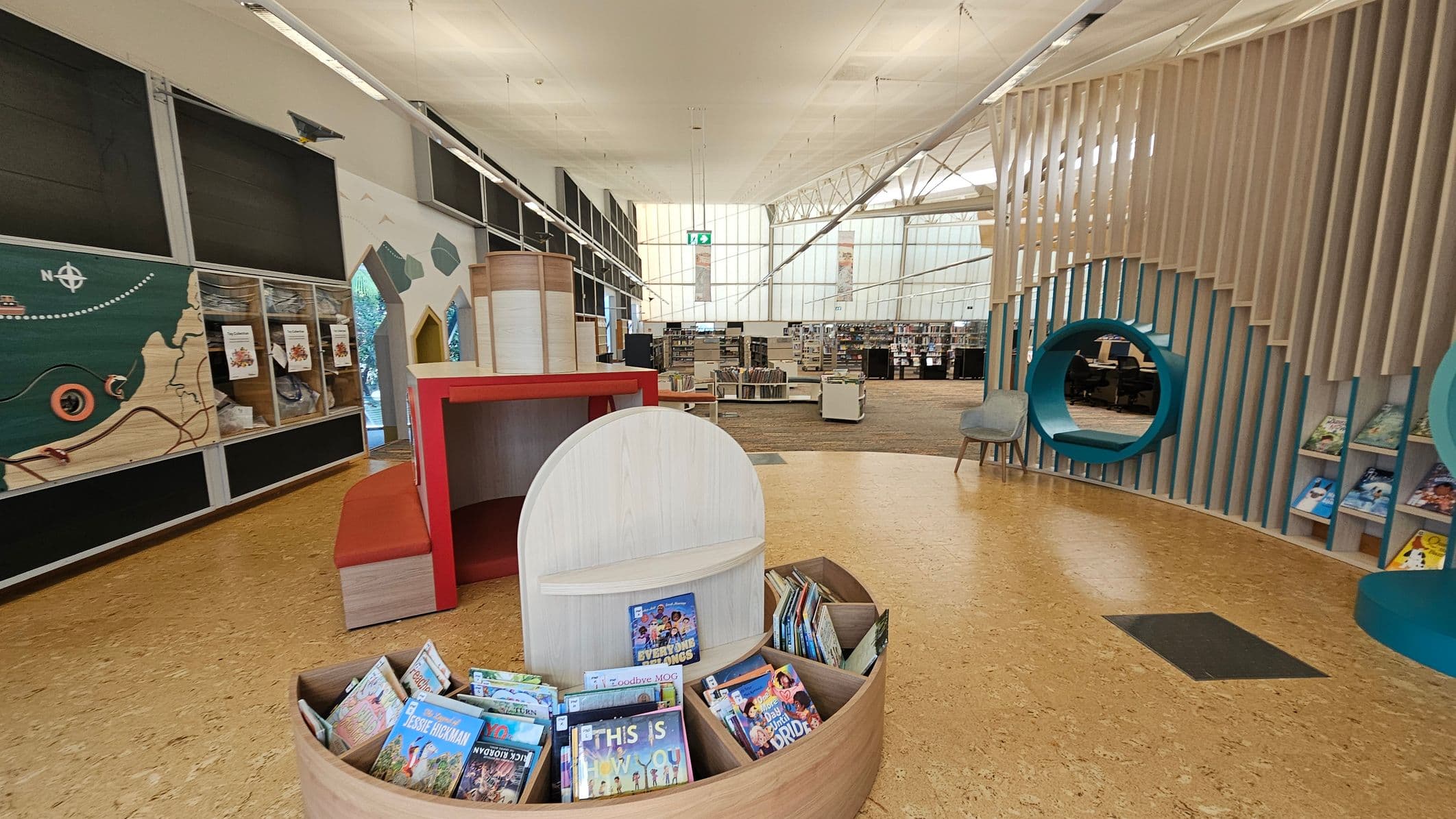 A modern library interior with a cork floor. On the left, a wall features a map design and display cases. In the centre, a circular wooden book display holds children's books. The background shows bookshelves and a large window with a grid pattern. On the right, a seating area with a circular cut-out in a slatted wall, accompanied by a chair. The ceiling is high with exposed beams and lighting fixtures.