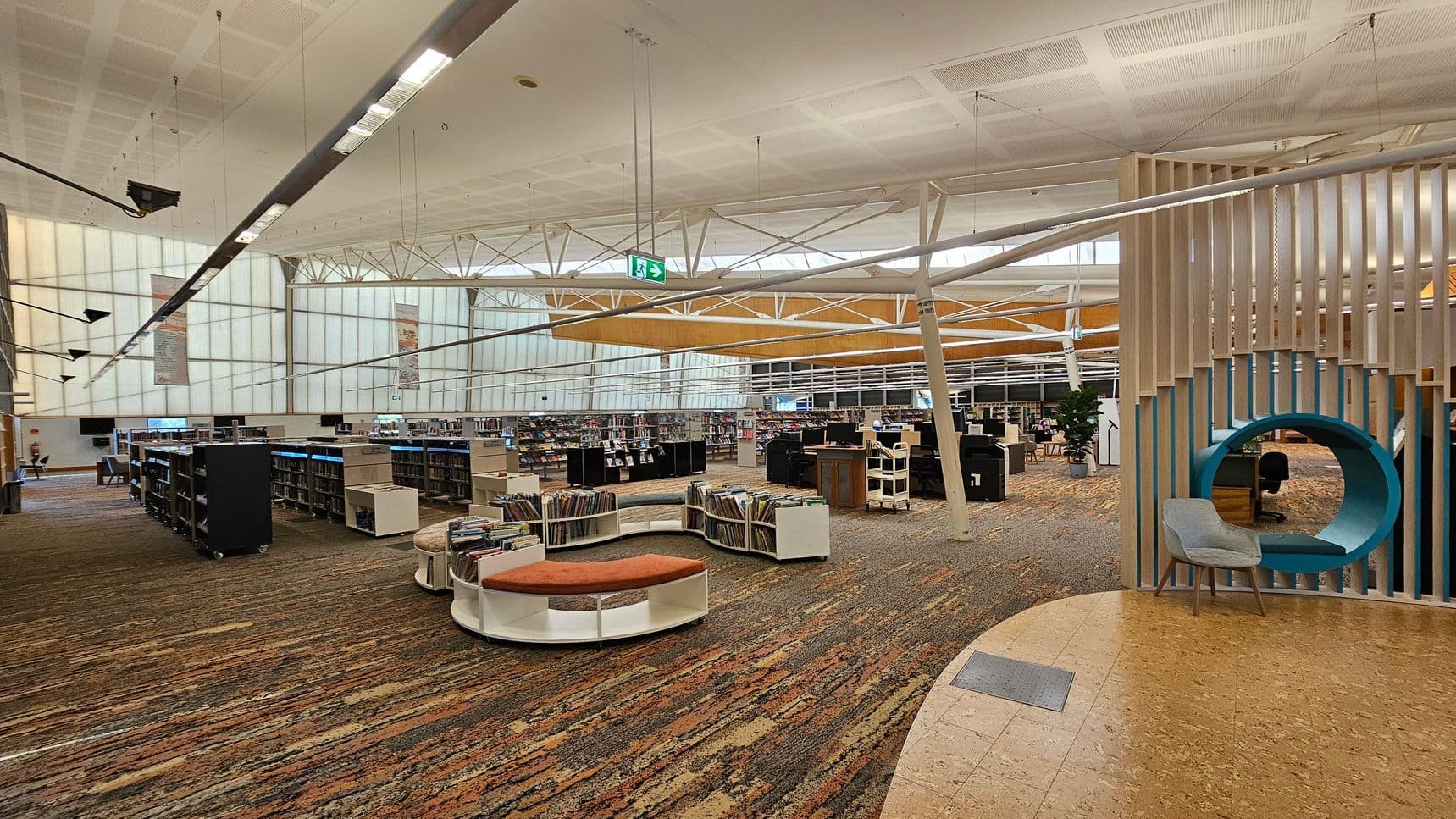 A spacious library interior with a high, white ceiling featuring exposed beams and hanging lights. On the left, there are rows of bookshelves filled with books. In the centre, a circular seating area with orange cushions is surrounded by more bookshelves. On the right, a unique seating nook with a blue circular frame and a grey chair is integrated into a wooden slat partition. The floor is covered with a patterned carpet, and a section of cork flooring is visible in the foreground.