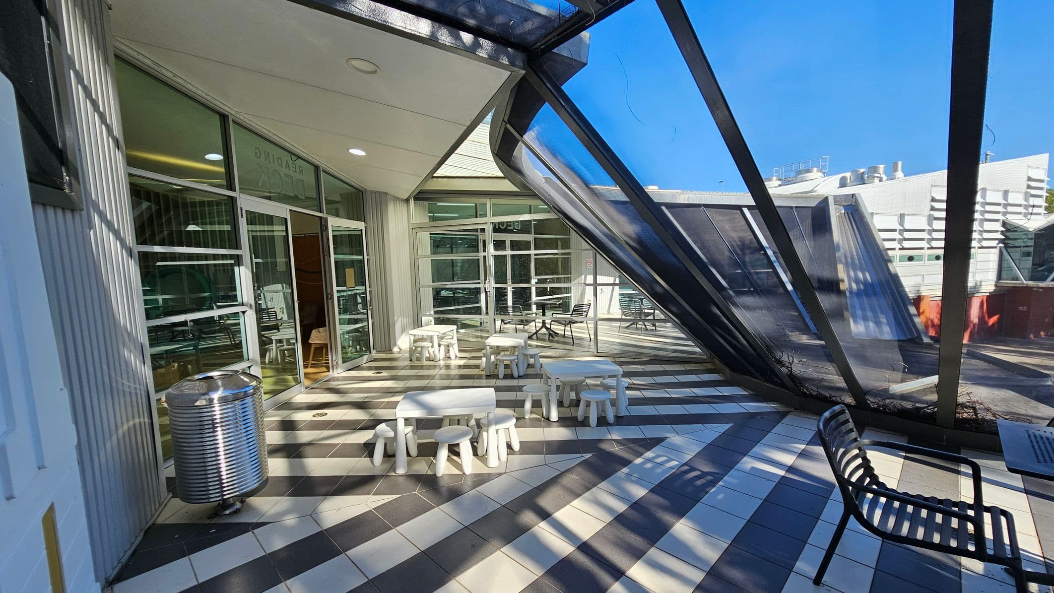 A modern indoor space with a slanted glass wall on the right, allowing natural light to flood in. The floor is tiled in a black and white checker pattern. In the centre, there are small white tables and stools arranged in a casual seating area. On the left, a metal bin is placed near glass doors leading to another room. A black metal bench is positioned on the right side. The ceiling is white with recessed lighting.