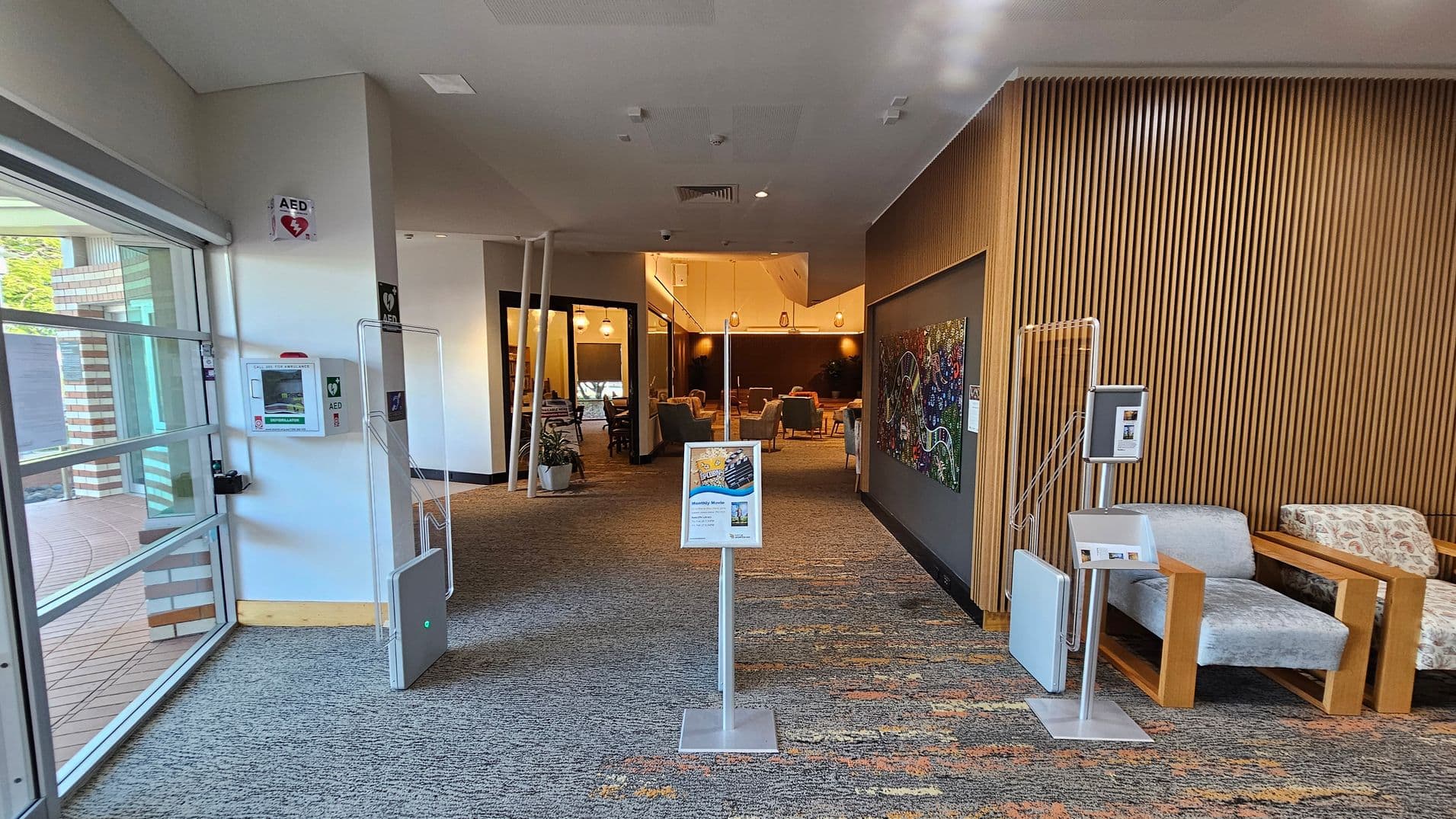 On the left, a glass wall with a view of the outside and an AED mounted on the wall. In the centre, a stand with a sign and a view into a seating area with tables and chairs. The right side features a wooden slatted wall with a colourful artwork and two cushioned chairs with wooden frames. The floor is carpeted with a multicoloured pattern, and the ceiling is white with recessed lighting.