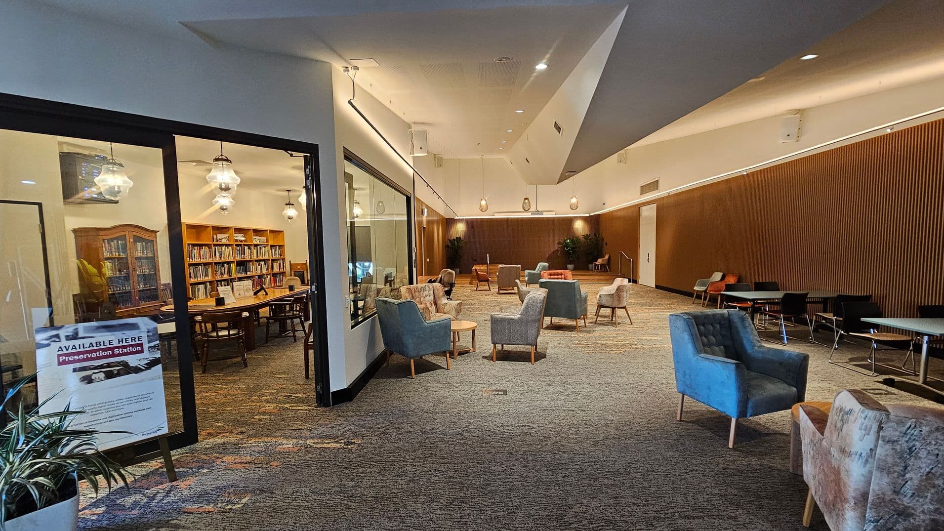 A spacious interior with a library room on the left, visible through a glass wall. The library has wooden bookshelves filled with books and a large table with chairs. A sign outside reads "Available Here Preservation Station." The main area has a variety of seating, including armchairs and small tables, on a patterned carpet. The ceiling is high with modern lighting, and the far wall is wooden with plants placed at intervals.