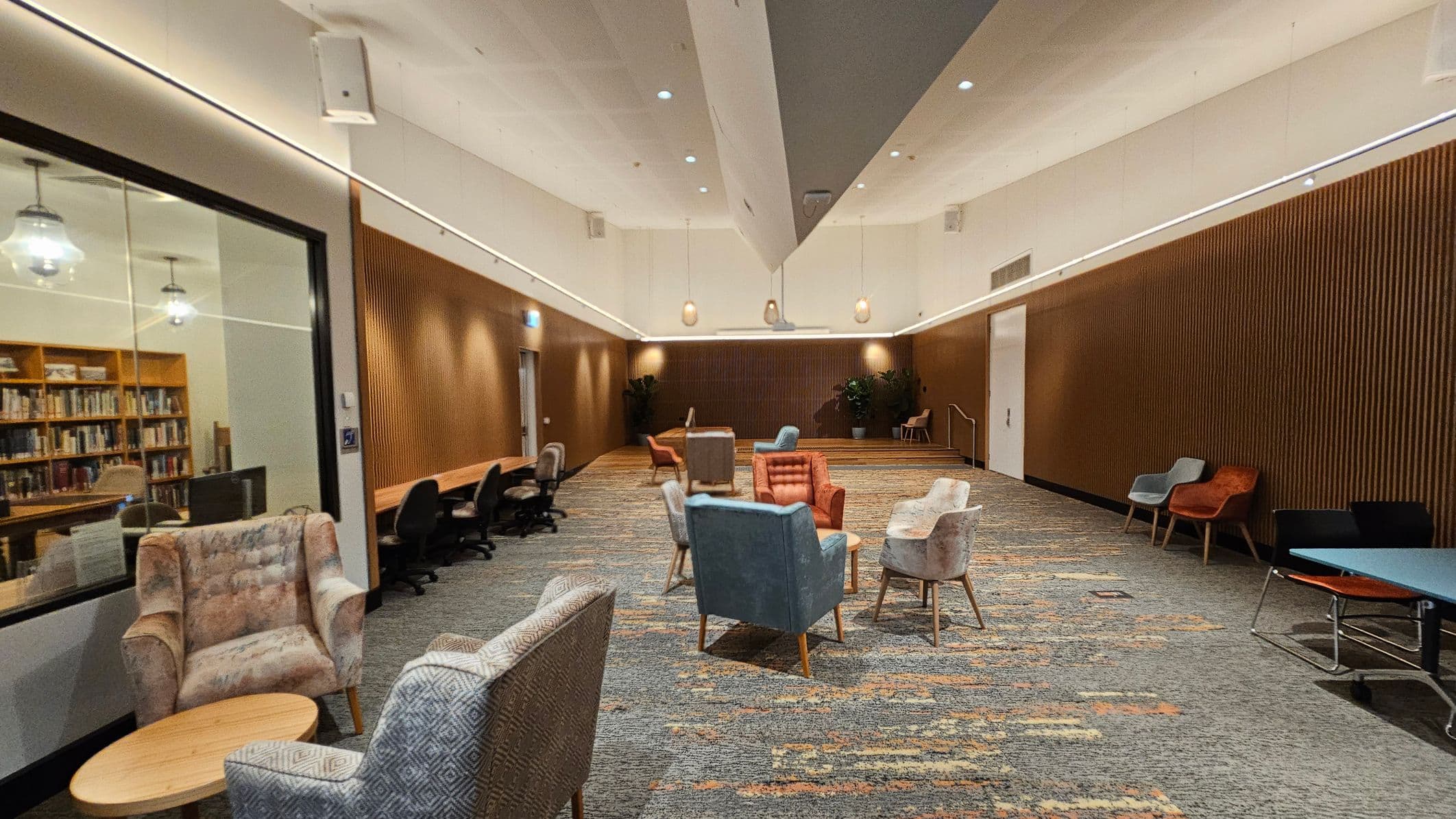 A spacious room with a high ceiling and modern lighting. On the left, there's a glass wall revealing a library with bookshelves. In the centre, a cluster of colourful armchairs surrounds small tables on a patterned carpet. The right side features a wooden panelled wall with additional seating, including chairs and a table. The floor is carpeted with a mix of green and orange tones, and there are potted plants in the background.