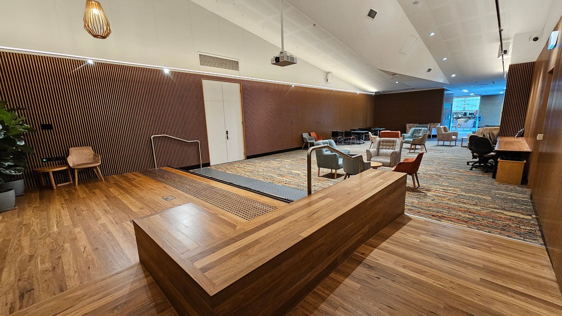 A modern interior space with a wooden floor and a slatted wooden wall on the left. A small wooden chair and table are near the wall. A ramp with metal railings leads to a carpeted area with various chairs and tables. A wooden counter is in the foreground. The ceiling is white with recessed lighting and a hanging light fixture. In the background, large windows allow natural light to enter, and there is a desk with an office chair on the right.