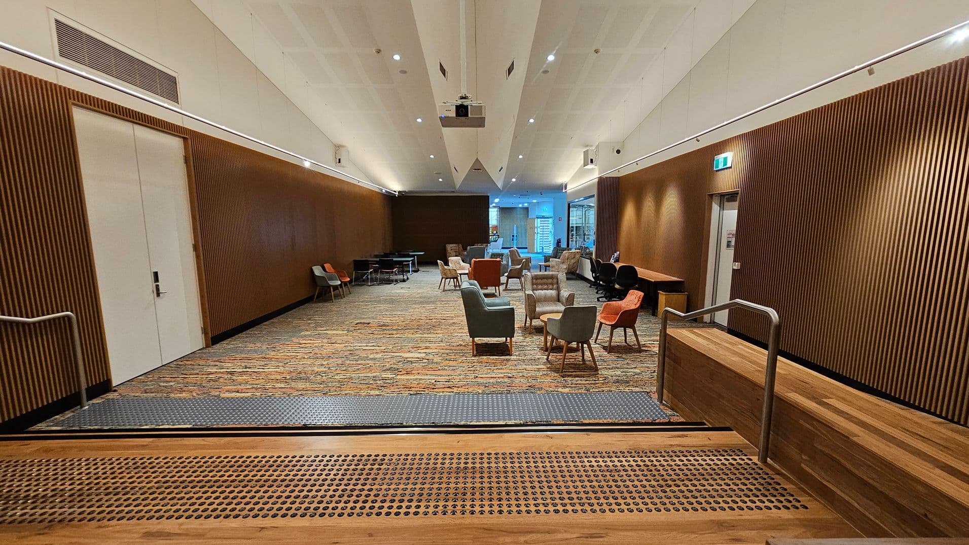 A spacious room with a patterned carpet floor and wooden panelled walls. In the centre, there are various chairs and small tables arranged in clusters. On the left, a large white door is set into the wall. On the right, a wooden desk and more seating are visible. The ceiling is white with recessed lighting and a projector. At the foreground, there are steps with a metal handrail leading down into the room.