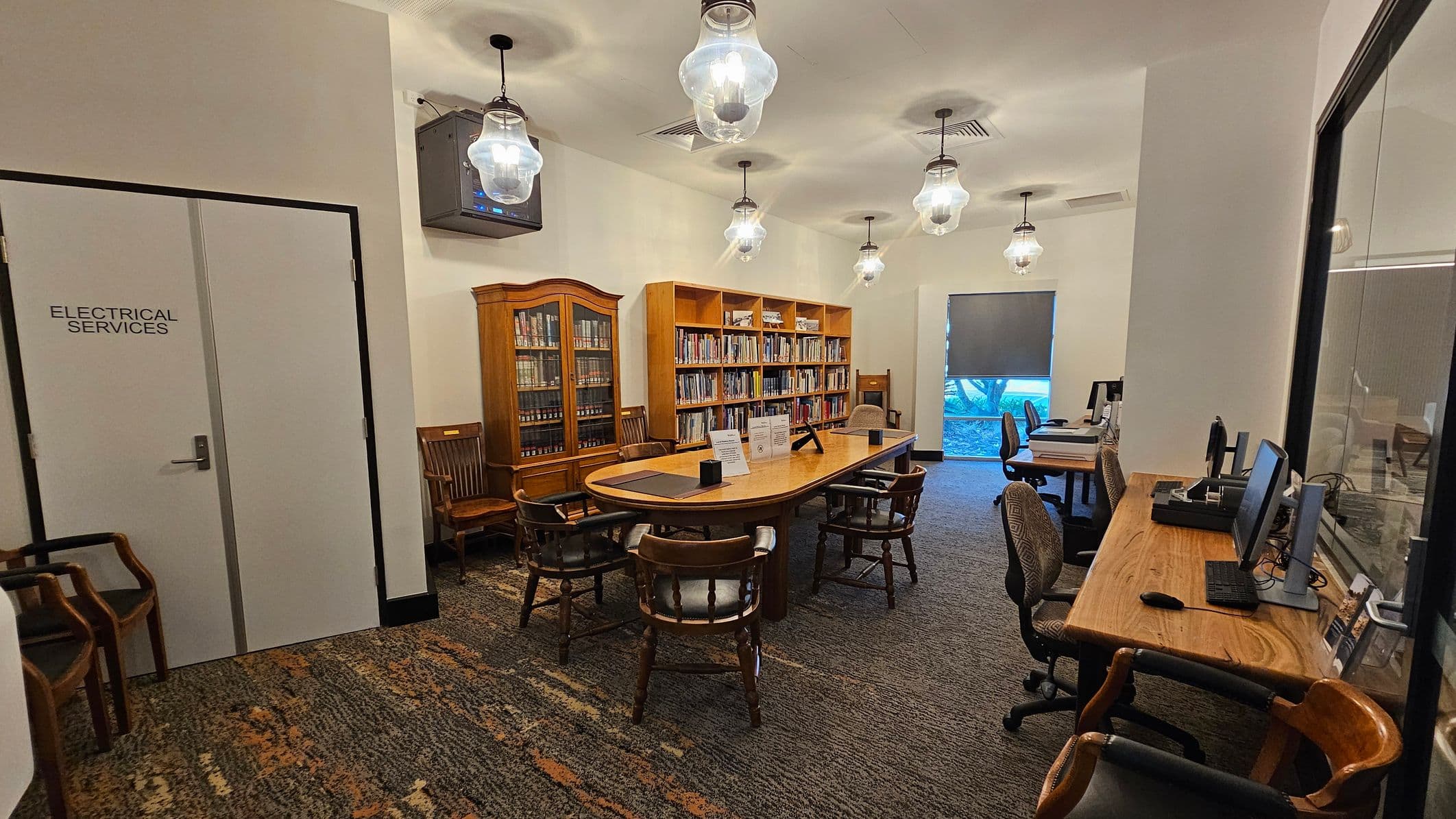 A library room with a carpeted floor. On the left, a door labelled "Electrical Services" and several wooden chairs. In the centre, a long wooden table surrounded by chairs, with bookshelves filled with books behind it. On the right, a wooden desk with computers and office chairs. The ceiling features several hanging light fixtures. A window with a partially drawn blind is in the background.
