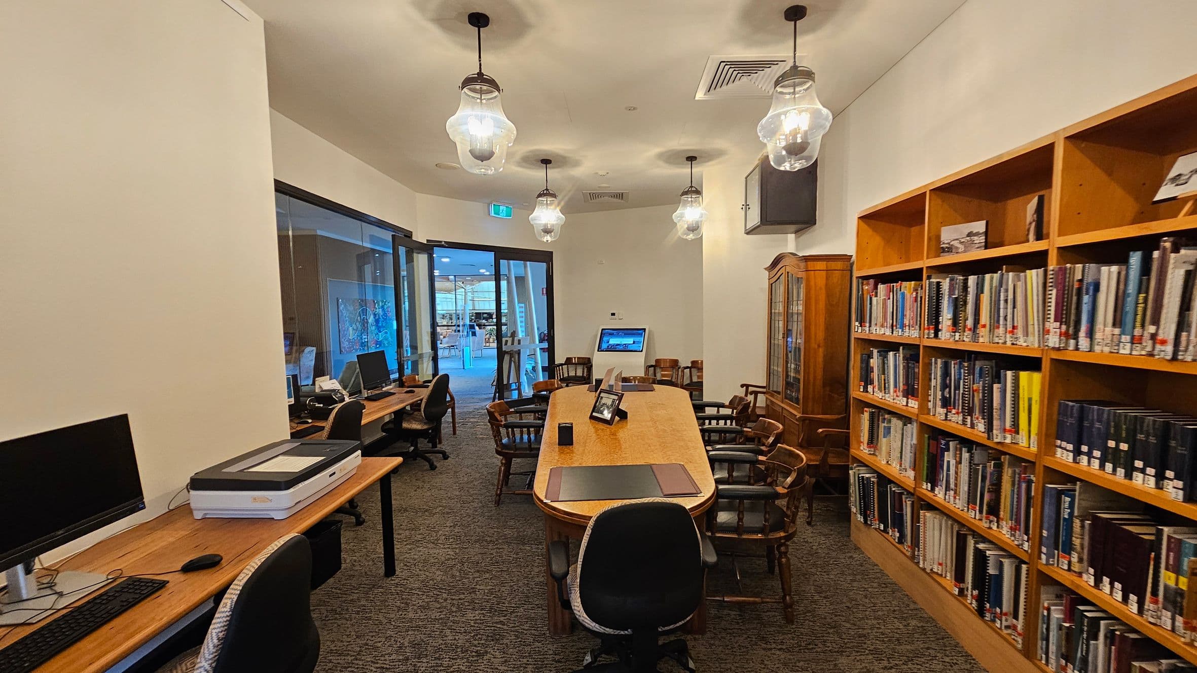 A library room with a long wooden table in the centre surrounded by wooden chairs. On the right, there are bookshelves filled with books. On the left, a row of desks with computers and a scanner. The floor is carpeted, and the ceiling has several hanging light fixtures. At the back, a glass door leads to another area with visible artwork and more seating.