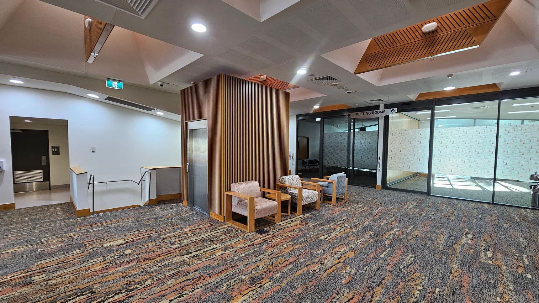 A modern interior space with a patterned carpet covering the floor. On the left, there's a staircase with a metal railing and a door. In the centre, a wooden panelled column houses a lift, with three cushioned chairs in front. On the right, a glass wall with a sign above reading "Meeting Rooms" leads to a room with frosted glass. The ceiling features recessed lighting and wooden accents.