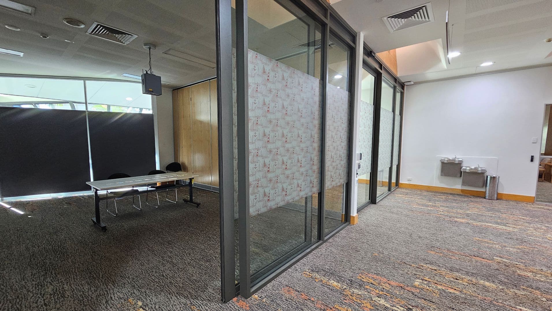 On the left, there is a room with a long table and several chairs, partially visible through a glass partition. The glass has a patterned design. The floor is carpeted with a mix of grey and brown tones. On the right, there are two water fountains mounted on a white wall, with a silver bin nearby. The ceiling has recessed lighting and air vents.