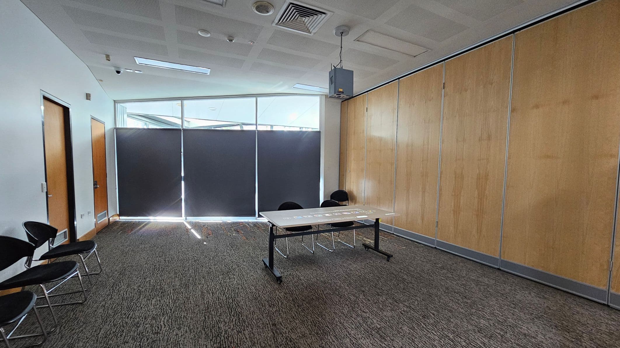A room with a grey carpeted floor and a white ceiling featuring recessed lighting and vents. On the left, there are two wooden doors and a row of black chairs with metal legs. The centre has a table with two chairs behind it. The right side has a wall with light wooden panels. The back wall has large windows covered with dark blinds, allowing some light to filter through. A projector is suspended from the ceiling.