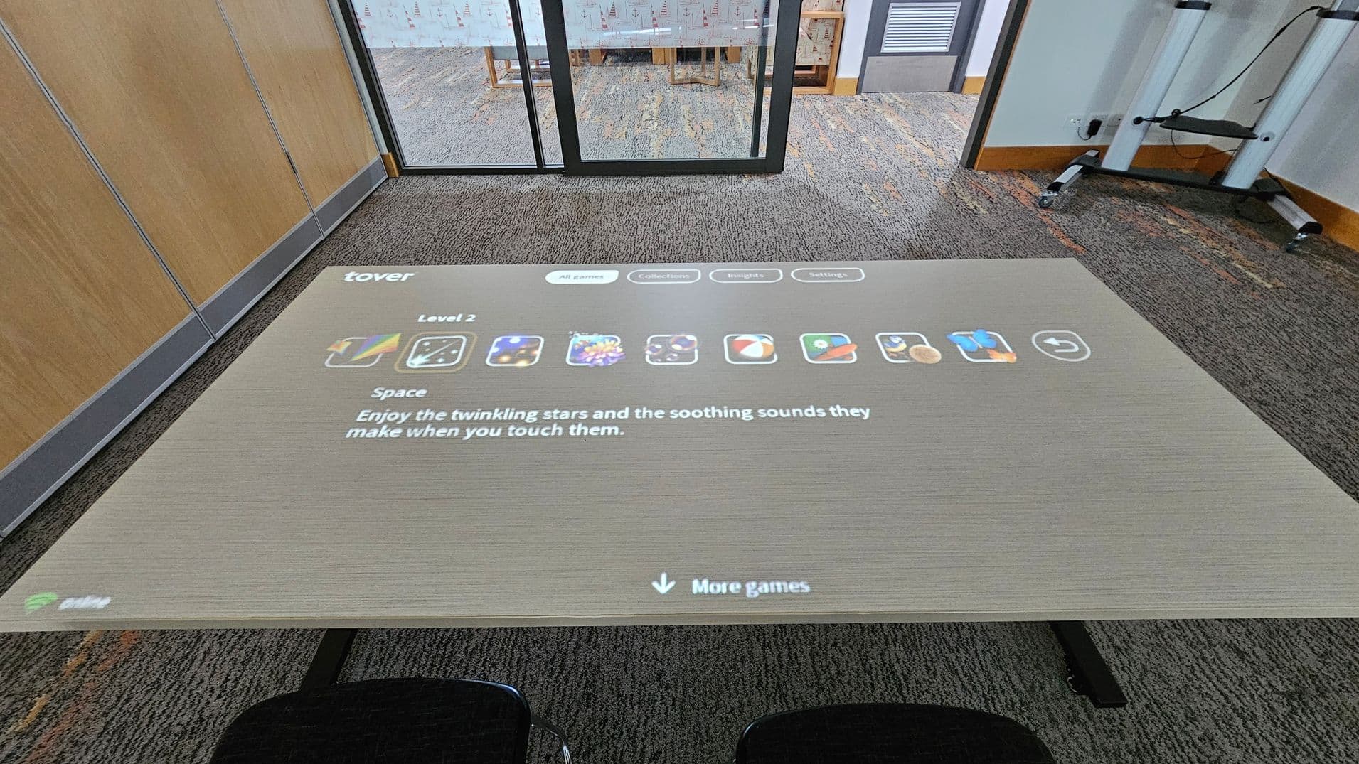A digital interface is projected onto a table, displaying a game menu with icons and text. The text reads, "Enjoy the twinkling stars and the soothing sounds they make when you touch them." In the background, there is a glass partition with a view into another room, which has a carpeted floor and a wooden chair. The floor is carpeted with a pattern, and there are wooden panels on the left wall. Two black chairs are partially visible in the foreground.