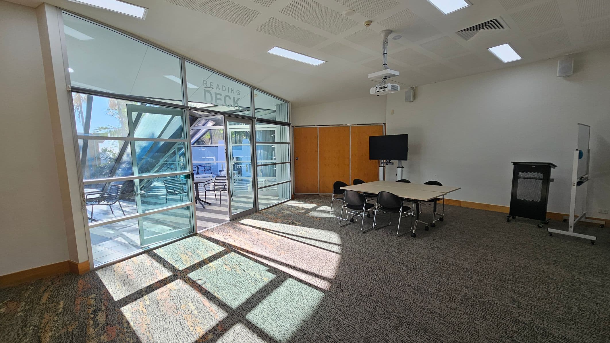 A room with a carpeted floor and a ceiling featuring recessed lighting and a projector. On the right, there's a table with several chairs, a TV on a stand, and a lectern. A whiteboard is also visible. On the left, large glass doors lead to an outdoor area with tables and chairs, labelled "Reading Deck." Sunlight streams through the glass, casting shadows on the floor.