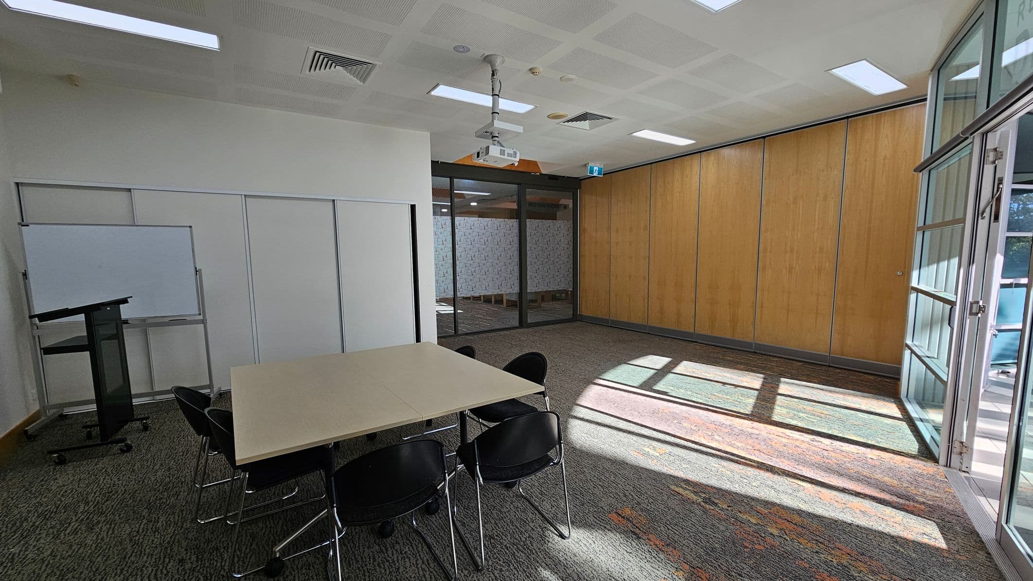 A conference room with a light-coloured table surrounded by black chairs on the left. A whiteboard and a lectern are positioned against the left wall. The right side features large wooden panels and glass doors, allowing natural light to stream in, casting shadows on the carpeted floor. The ceiling has recessed lighting and a mounted projector.