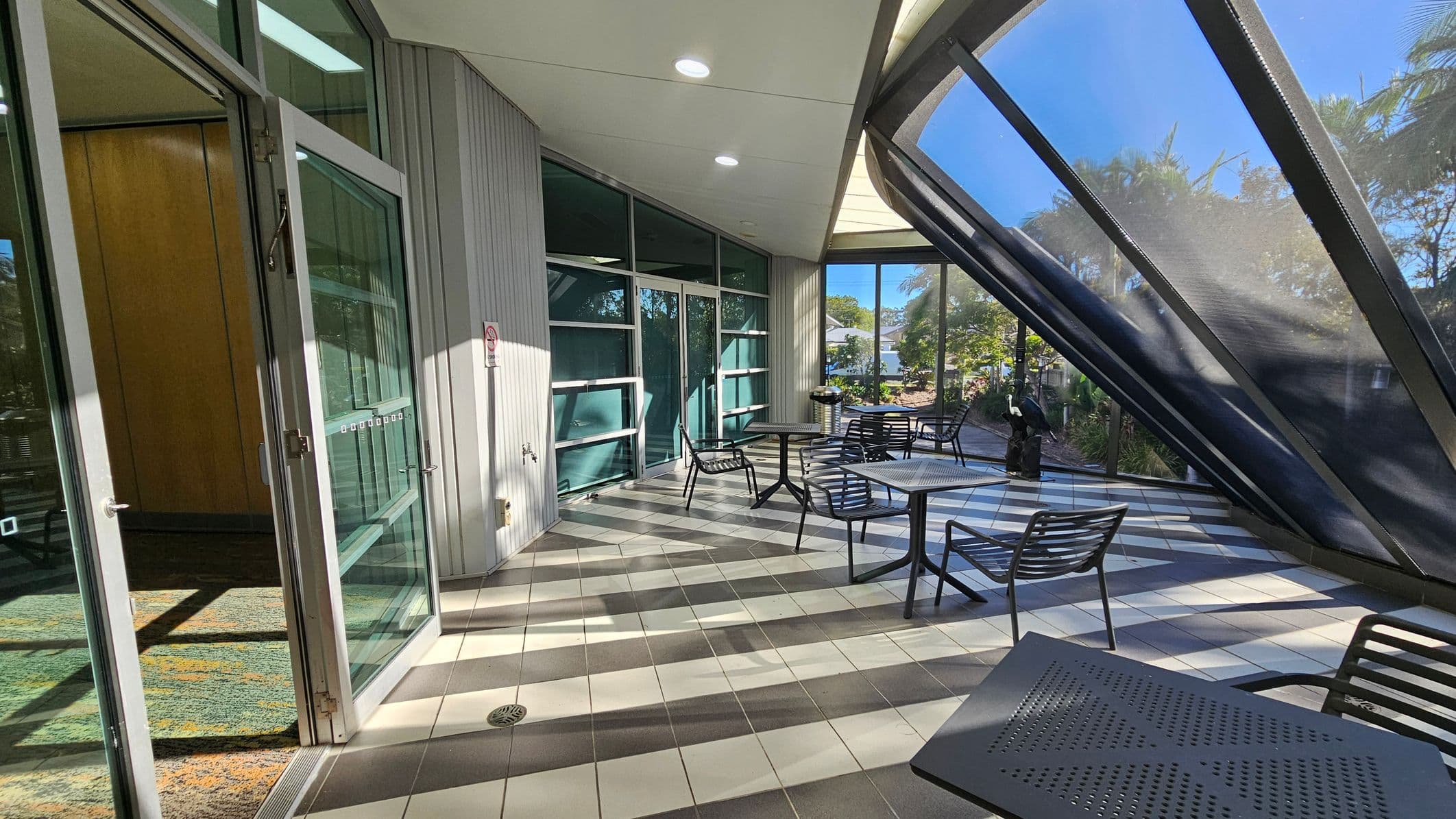 A modern indoor space with a slanted glass wall on the right, allowing natural light to flood in. The floor is tiled in a checkered pattern of grey and white. On the left, there's a glass door leading to an interior area with a patterned carpet. Several black metal tables and chairs are arranged throughout the space. In the background, greenery is visible through the glass, and a small sculpture is near the window. The ceiling is smooth with recessed lighting.