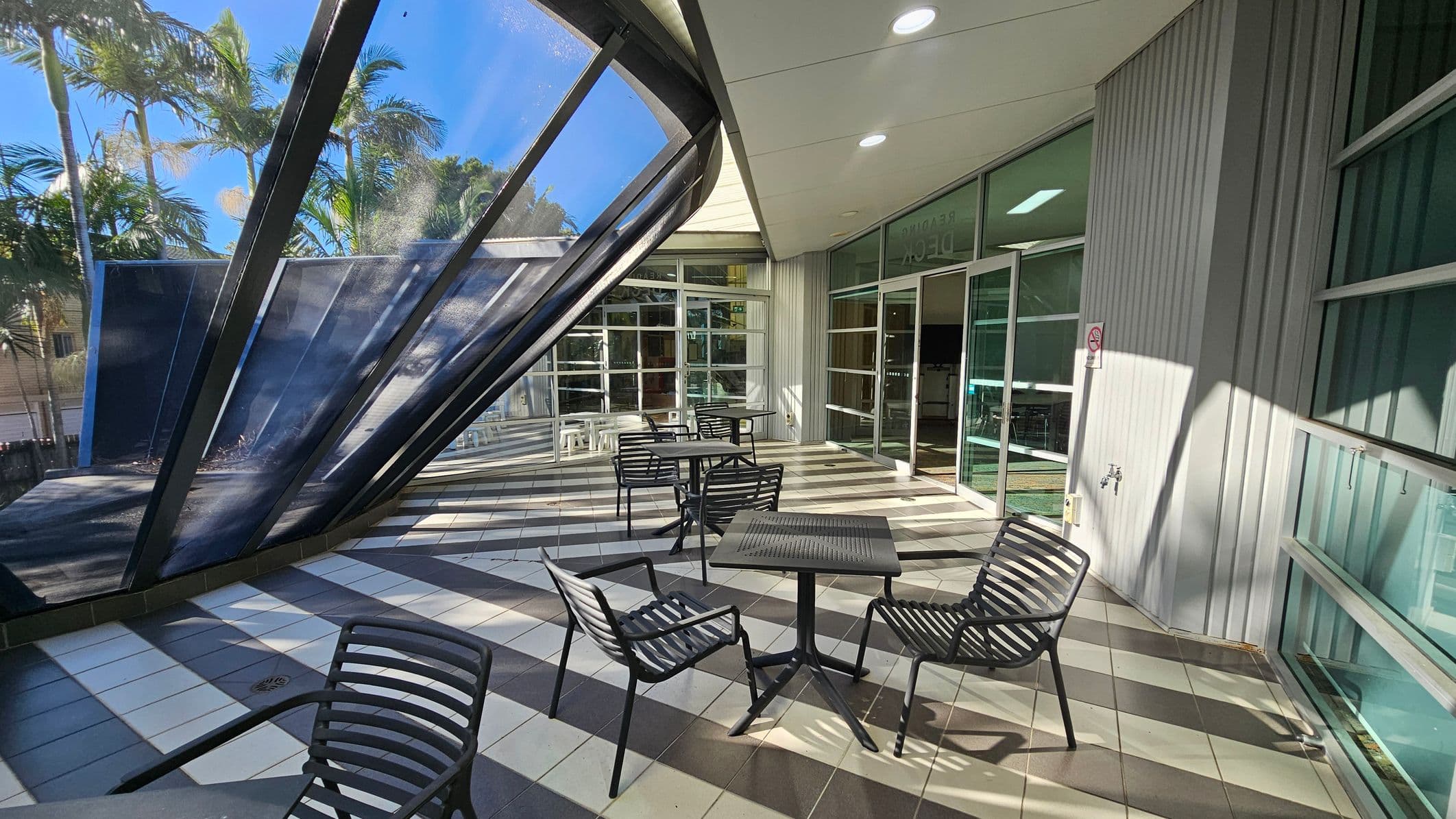 Outdoor seating area with black metal tables and chairs on a tiled floor featuring a striped pattern. On the left, a large, slanted glass structure with metal frames provides partial shade. In the background, tall palm trees are visible. The right side has a building with large glass doors and windows, and a small wall-mounted tap. The ceiling is white with recessed lighting.