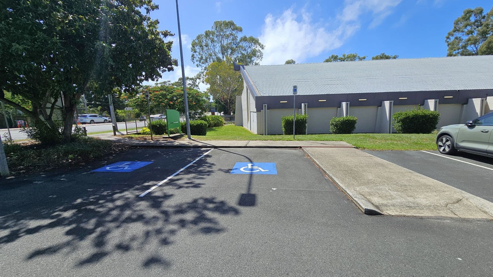 A car park with two marked disabled parking spaces on the left, featuring blue wheelchair symbols. A green car is parked on the right. A large tree provides shade on the left, and a building with a grey roof and small bushes is in the background. A green sign and a street with a white car are visible in the distance. The ground is asphalt, and there is a concrete ramp between the parking spaces and the building.