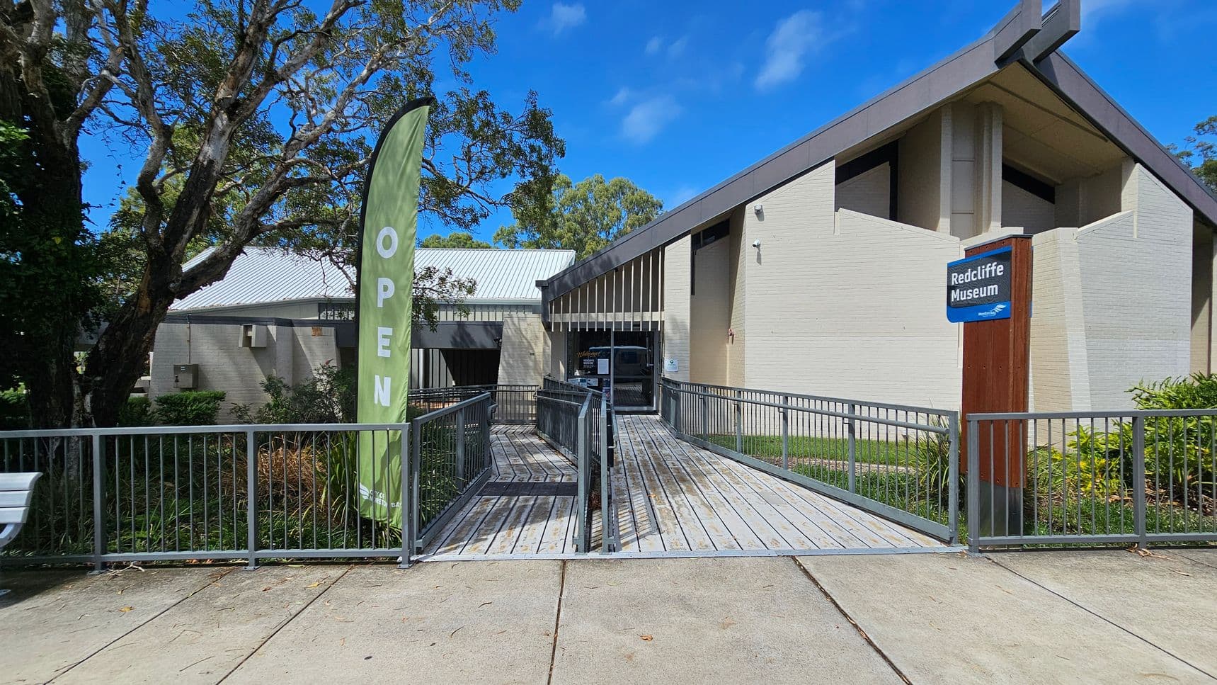 A modern building with a sloped roof and light-coloured brick walls. In the foreground, a wooden ramp with metal railings leads to the entrance. A green flag with the word "OPEN" is on the left, next to a large tree. On the right, a sign reads "Redcliffe Museum" mounted on a wooden post. The ground is paved, and there are plants and shrubs around the building. The sky is clear and blue.
