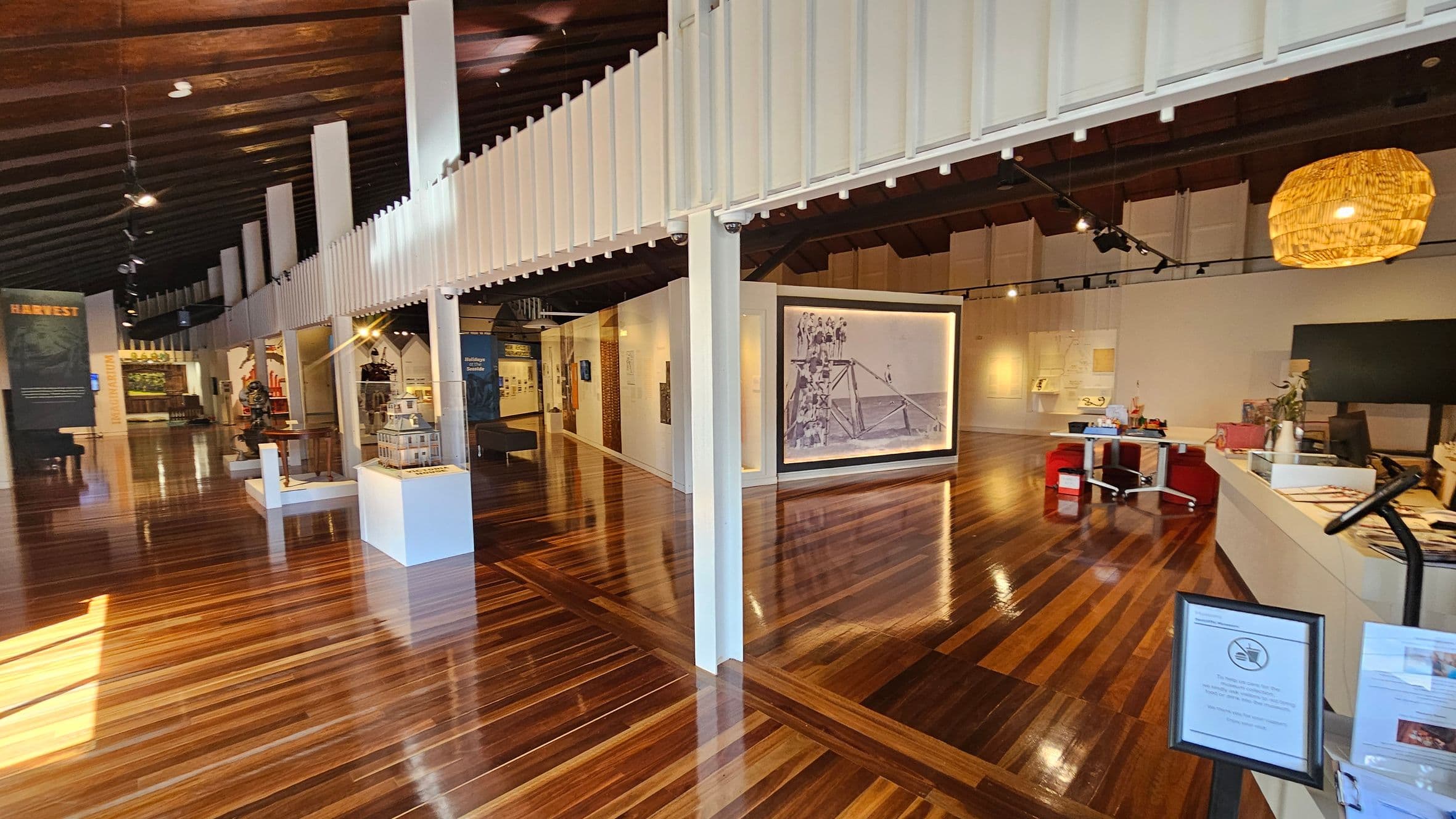 A spacious interior with polished wooden floors and a high, dark wooden ceiling. On the left, there are various exhibits, including a model house on a pedestal and a large display board. In the centre, a large photograph is mounted on a wall. To the right, a reception area with a counter, chairs, and a hanging light fixture. White pillars support a mezzanine with a railing above. The space is well-lit with track lighting.
