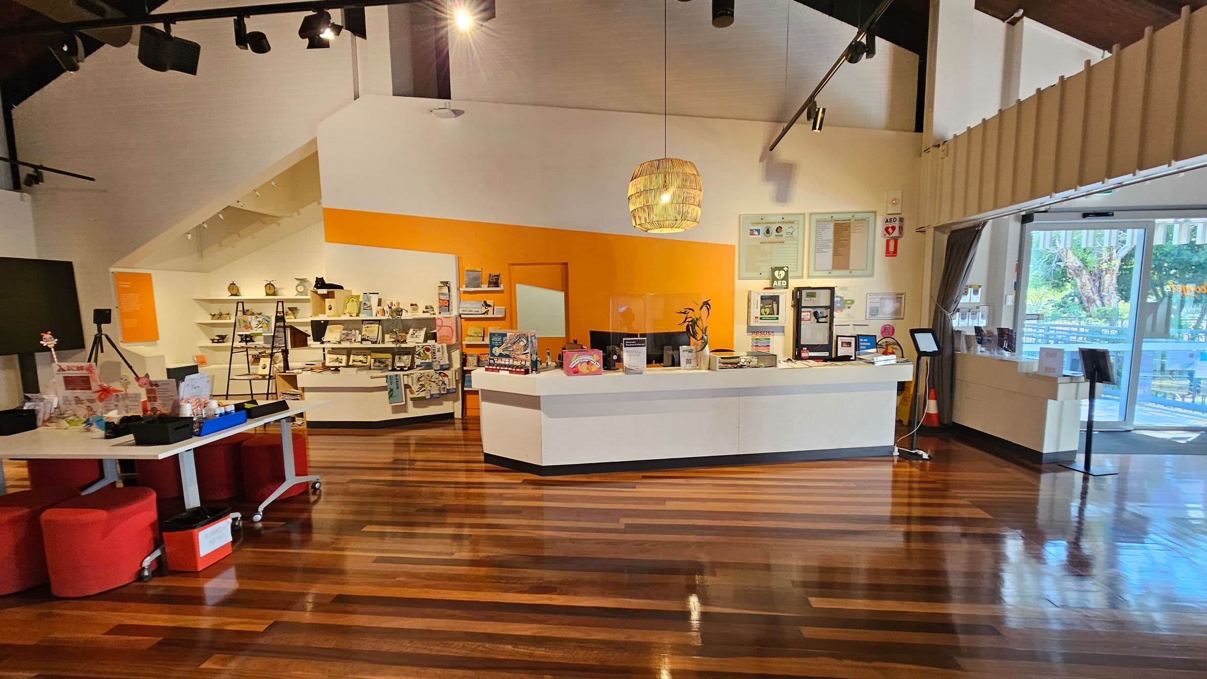 A spacious interior with a wooden floor and high ceiling. On the left, there are red stools and a table with various items. In the centre, a white reception desk is set against an orange wall, with shelves displaying books and merchandise. On the right, a glass door leads outside, and there are informational displays and a digital kiosk. A woven pendant light hangs above the desk.