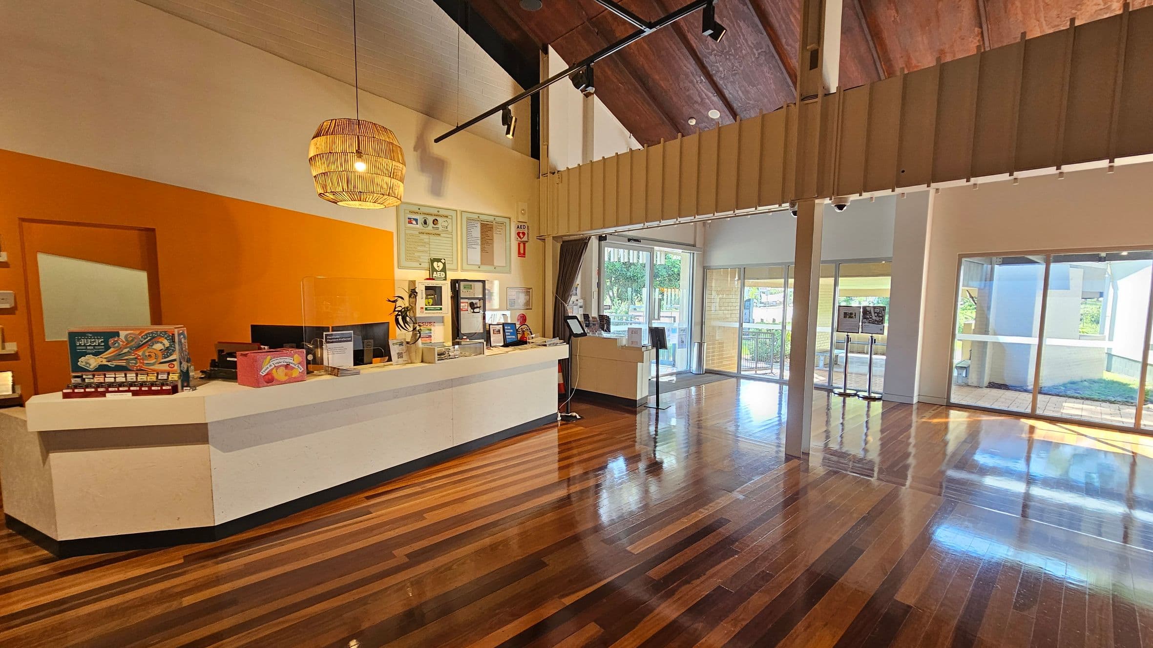 Reception area with a white and orange counter on the left, displaying brochures and a computer. A wicker pendant light hangs above. The floor is polished wood, reflecting the light. On the right, large glass doors and windows offer a view of the outside, with greenery visible. The ceiling is high with exposed wooden beams, and a track lighting system is installed. A small display stand is near the entrance.