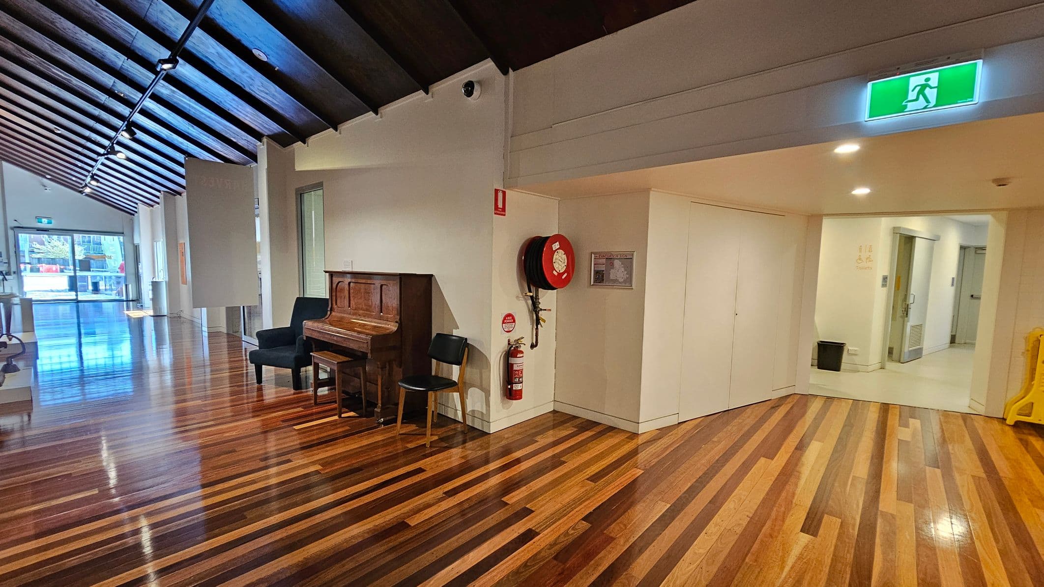 A hallway with polished wooden floors and a dark wooden ceiling with spotlights. On the left, there's a glass door leading outside. In the middle, a wooden piano with a black chair is placed against the wall. On the right, a fire hose reel and extinguisher are mounted on the wall near an emergency exit sign. The corridor leads to a lighter-coloured area with a metal door and a small black bin.