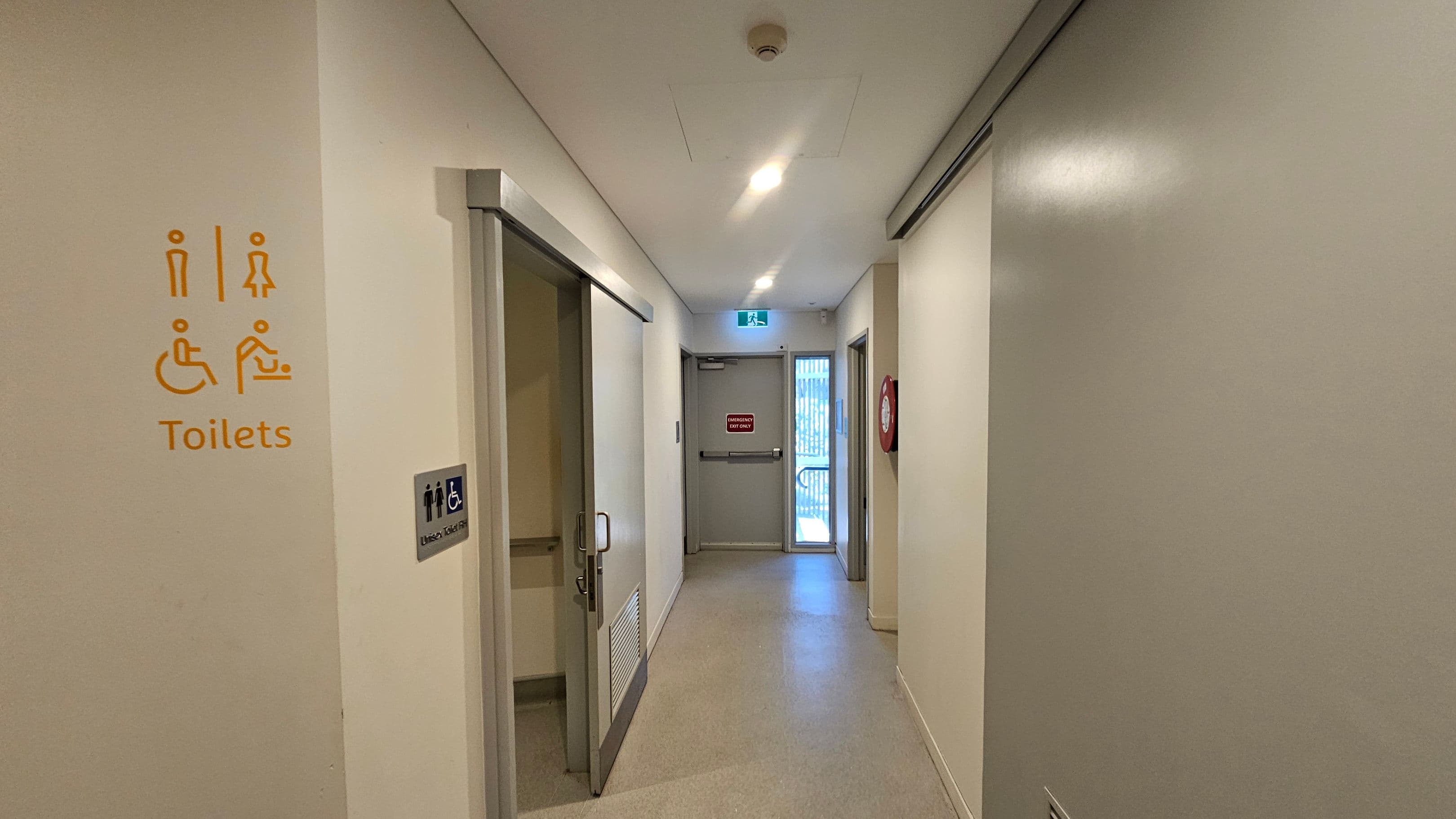 A corridor with light-coloured walls and a smooth floor. On the left, there is a partially open door with a sign indicating accessible toilets, including symbols for male, female, disabled, and baby changing facilities. The right wall is plain. At the end of the corridor, there is a door marked "Emergency Exit Only" with a lit exit sign above it. A fire hose reel is mounted on the right wall near the exit. The ceiling is fitted with recessed lighting.