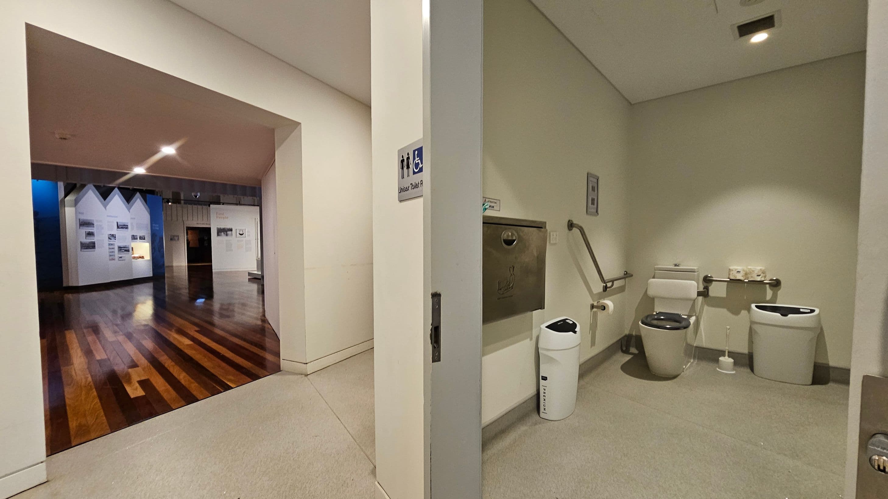 On the left, a hallway with polished wooden flooring leads to an exhibition area with white display panels featuring photographs and text. On the right, an accessible toilet room with a grey floor, a toilet with support rails, a sanitary bin, and a paper towel dispenser. The ceiling is plain with recessed lighting.