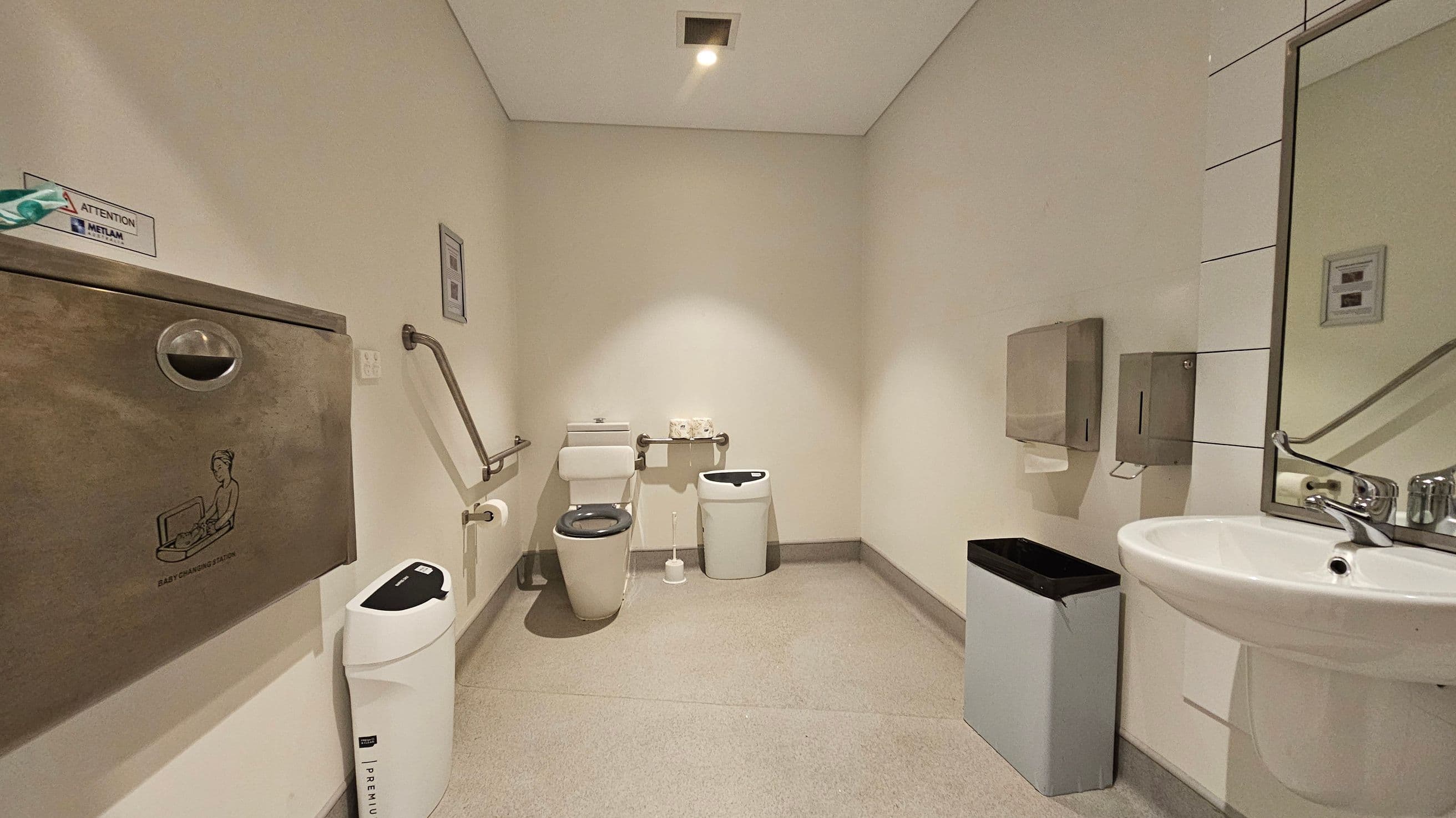 A public restroom with a baby changing station on the left wall, above a white bin. In the centre, a toilet with a support rail and a small bin nearby. On the right, a sink with a mirror above it, and a paper towel dispenser mounted on the wall. The floor is light grey, and the ceiling is white with a recessed light.