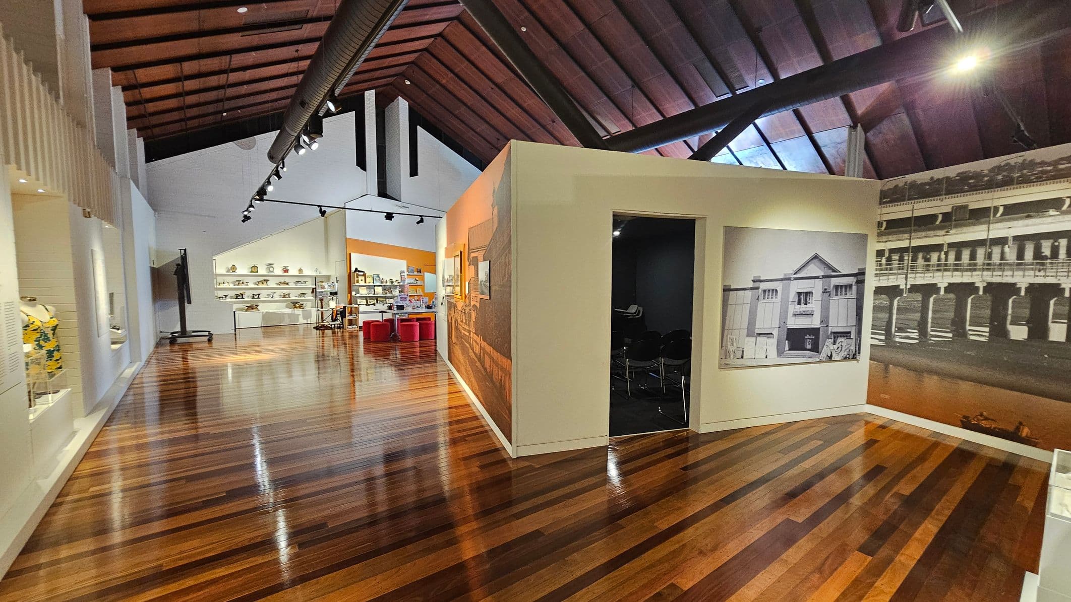 A museum interior with polished wooden floors and a high, wooden-beamed ceiling. On the left, there are display cases with various items, including clothing. In the centre, a white partition wall features black-and-white historical photographs. To the right, more photographs are displayed on the wall. In the background, shelves with pottery and other artefacts are visible, along with red seating and a bright orange wall. Overhead, black track lighting is installed.