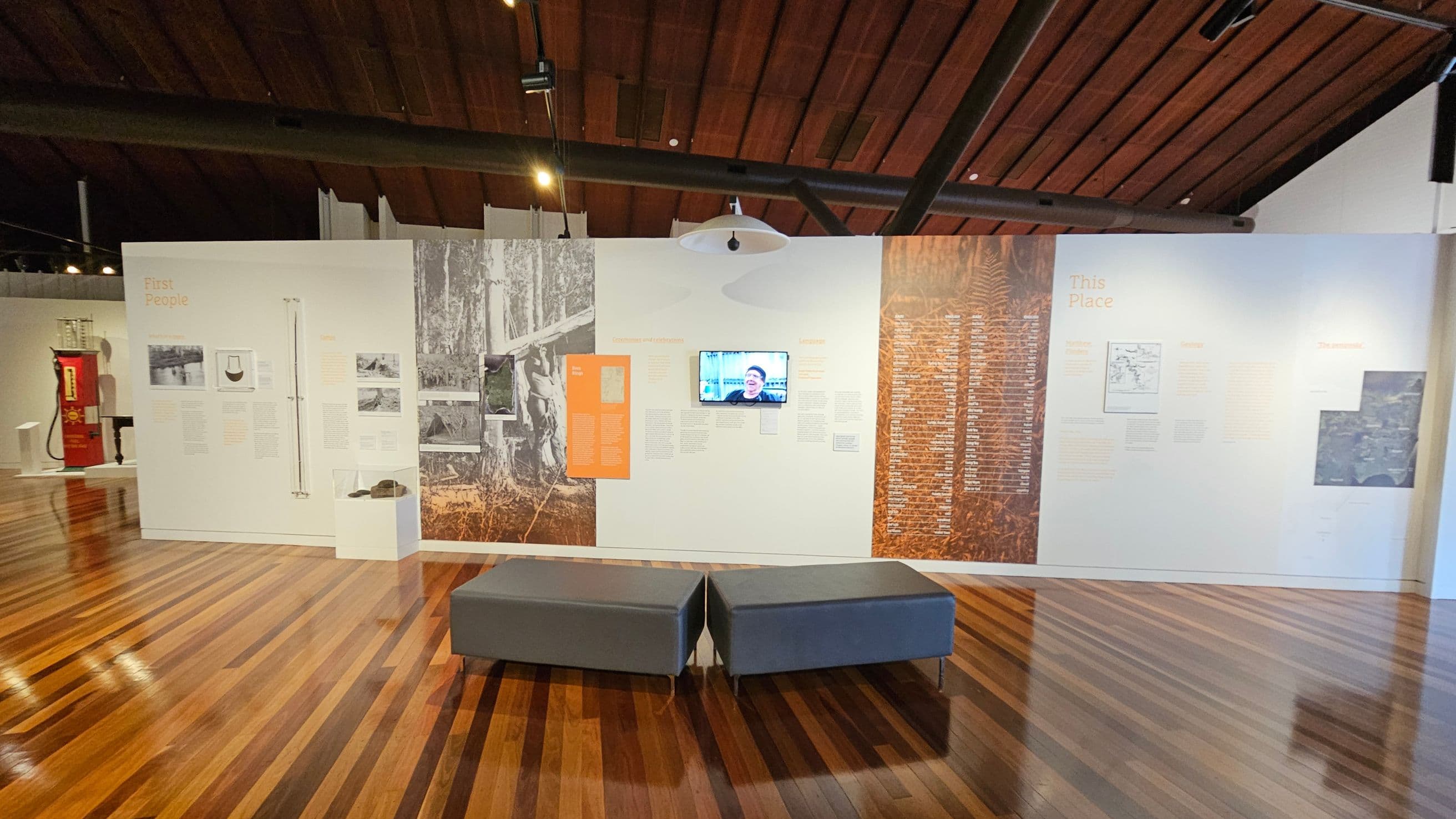 A museum exhibit with a wooden floor and a high, wooden ceiling. The back wall features informational panels with text and images, including a small screen displaying a person. Two grey benches are positioned in the foreground. On the left, a red vintage petrol pump is visible. The exhibit appears to focus on historical or cultural themes, with sections titled "First People" and "This Place."