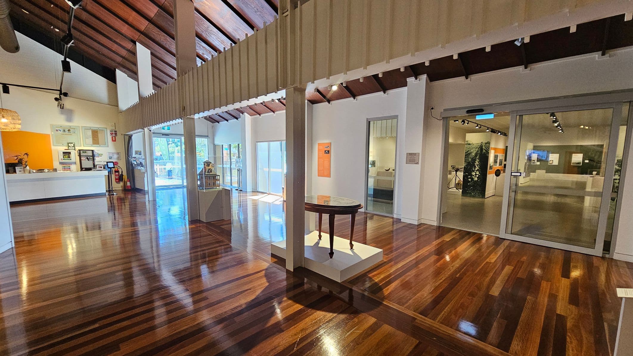A spacious interior with polished wooden floors and a high, wooden-beamed ceiling. On the left, a white reception desk with a bright orange wall behind it, displaying various framed items. In the centre, a small round table on a white platform and a model house on a pedestal. To the right, glass doors lead into another room with displays and exhibits. Large windows in the background allow natural light to flood the space.