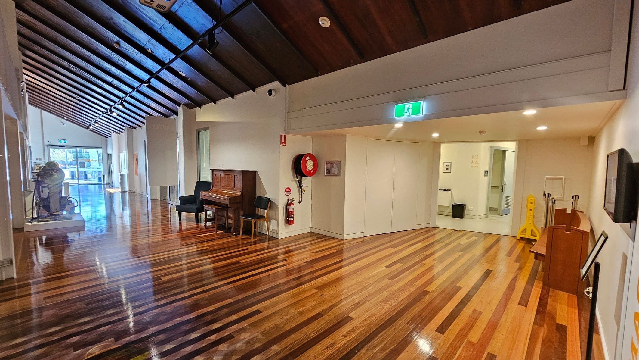 A hallway with polished wooden floors and a dark wooden ceiling with exposed beams. On the left, there's a piano with two chairs and a fire hose reel mounted on the wall. Further down, a large machine is displayed. On the right, a doorway leads to another room, with a yellow caution sign nearby. The walls are light-coloured, and there are overhead lights. An exit sign is visible above the doorway.