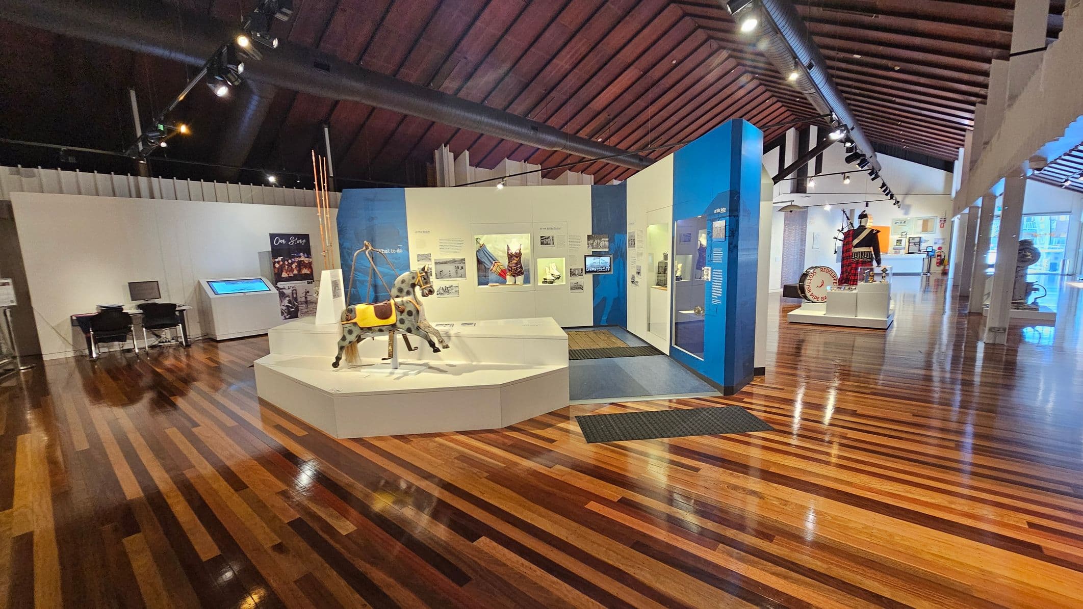 A museum interior with polished wooden floors and a high, dark wooden ceiling. On the left, there are two chairs and a desk with a computer. In the centre, a display features a carousel horse and various artefacts on white platforms and blue panels. On the right, a mannequin dressed in traditional attire stands next to a large drum. The background includes more exhibits and large windows allowing natural light. Track lighting is visible on the ceiling.